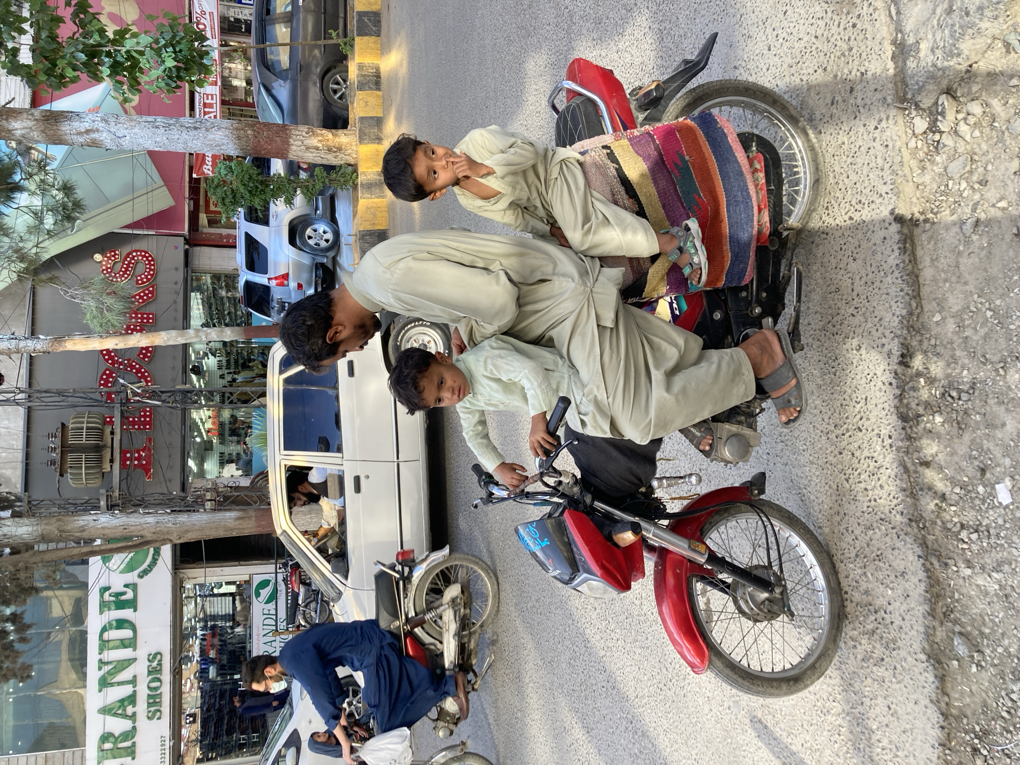 This is an outdoor, vertical-format photograph capturing a lively street scene in Quetta City Tehsil, Pakistan, under bright daylight. The central focus is a man and two young boys on or near a red motorcycle. The man, wearing a light-colored shalwar kameez, leans over towards a younger boy, also in a similar traditional outfit, who is positioned near the motorcycle's handlebars. Another boy, dressed similarly, sits on the motorcycle's seat, which is adorned with a colorful, striped woven fabric. This boy looks directly at the camera with a curious or playful expression, his finger near his mouth.
In the background and surrounding areas, various vehicles are visible, including a white sedan, a silver SUV, and a black SUV parked along the street. Another person, wearing a blue outfit, is partially visible on a different motorcycle or vehicle further back. The street features a curb painted with yellow and black stripes. Buildings line the thoroughfare, housing storefronts and businesses.
Visible text from the storefronts includes "GRANDE SHOES" and "SHOES" multiple times, suggesting a shoe store. Other snippets like "Ba" (possibly part of "Bata"), "ALE" (likely "SALE"), "0%OFF" (indicating a discount), and "ve collection" are discernible, along with a phone number "3322927." Trees with green foliage are scattered along the roadside, adding to the urban landscape. The overall atmosphere depicts a typical bustling street environment in a South Asian city.