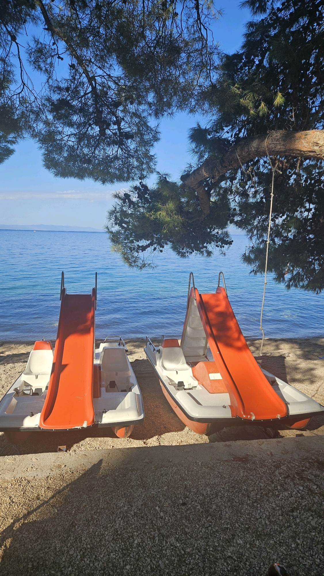Two pedal boats with orange slides are parked on a pebble beach next to the sea. The water is clear and blue, with a hazy coastline in the distance. A large pine tree with dark green needles is prominent in the foreground, casting shadows on the beach. The tree's branches extend over the scene, and a rope hangs from one branch, suggesting a swing or some other amenity. The sky is a clear, bright blue. The scene appears to be in Općina Tučepi, Croatia, a coastal town known for its beaches and natural beauty.