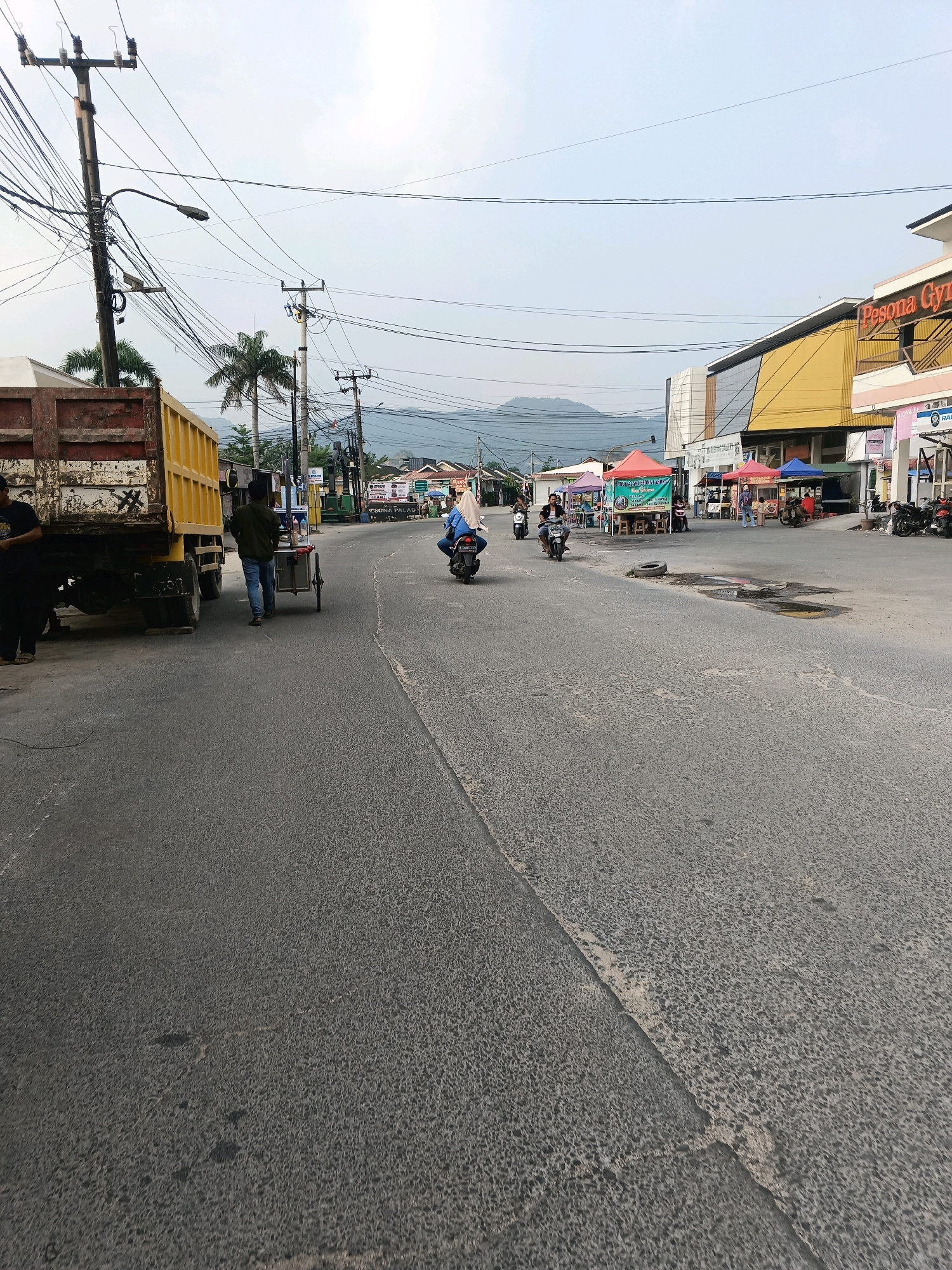 The image depicts a street scene in Klapanunggal, Indonesia, under an overcast sky. A yellow dump truck is parked on the left side of the asphalt road. A man in a dark shirt stands beside the truck. Further down the road, another man walks behind a cart. Several motorcycles are present, with one ridden by a person wearing a blue headscarf and another by a person in a white shirt. Along the right side of the road, there are several market stalls with colorful tents, and buildings, including one with a yellow facade and the signage "Pesona Gym". Mountains are visible in the background, and utility poles with numerous wires are scattered throughout the scene. The road surface shows some wear and tear, with visible patches and a puddle.