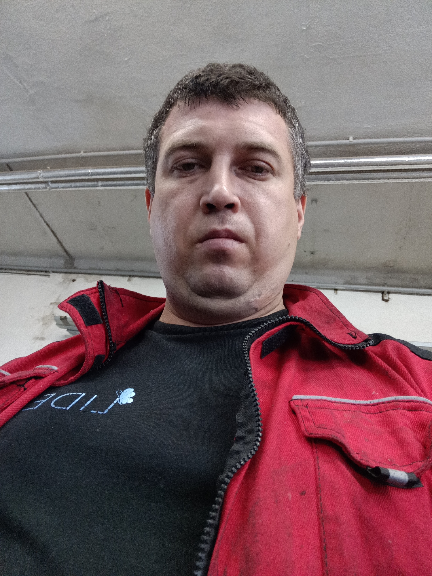 A low-angle portrait features a man looking directly into the camera with a neutral, serious expression. He appears middle-aged, with short, dark hair showing some grey at the temples. He is wearing a black t-shirt partially visible under an open, red work jacket or coverall. The jacket has a prominent black zipper and black velcro patches on the collar. On the wearer's right side, a flap pocket with a grey reflective strip is visible. The red fabric of the jacket appears somewhat worn and soiled, suggesting a working environment. On the black t-shirt, the word "FIDE" is visible in light blue/white lettering, accompanied by a small, light blue symbol that resembles a clover or butterfly.
The background shows a textured, light-colored ceiling, likely concrete or plaster, with visible imperfections. Industrial-style pipes or conduits are mounted horizontally across the ceiling in the upper background. The overall setting suggests an indoor, possibly workshop or industrial environment. The lighting is artificial. The photograph was taken in Dalovy, Czechia.