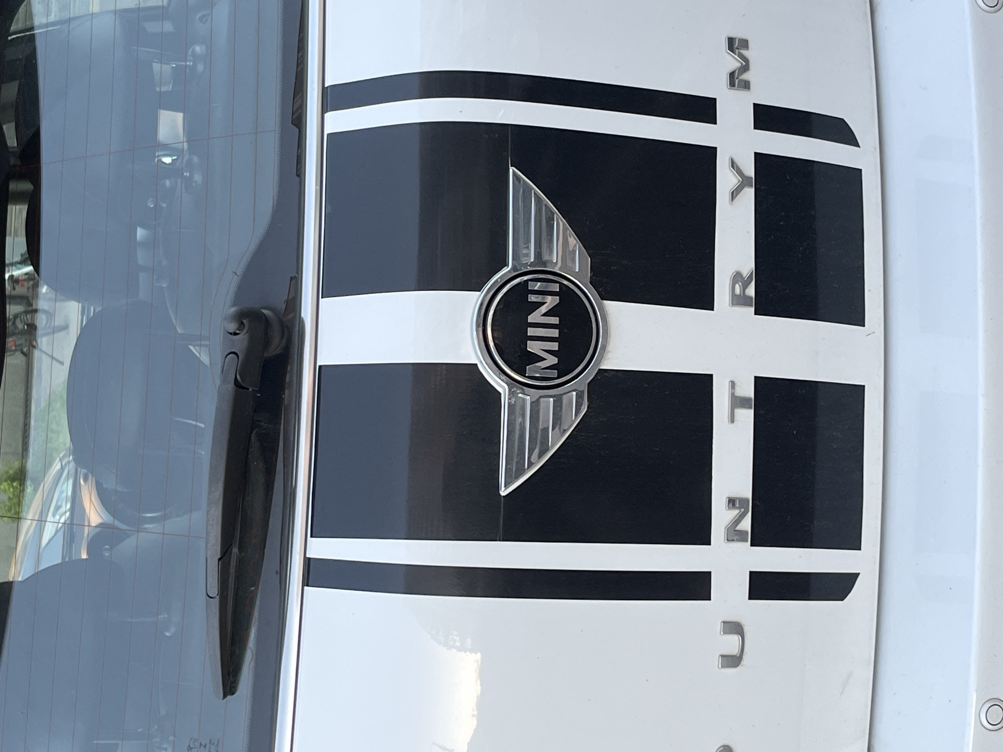 The image shows the rear of a white MINI Countryman car. The car has two black racing stripes running vertically down the center of the trunk. In the middle of the stripes is the MINI logo, which consists of a circular emblem with "MINI" written inside and wings on either side. Below the MINI logo, the word "COUNTRYMAN" is spelled out in silver lettering across the bottom of the trunk, with the letters "C", "U", "N", "T", "R", "Y", and "M" visible. The car is parked outdoors, possibly in a city, as suggested by the reflection of buildings and trees in the car's rear window and the presence of a parking sensor on the bumper. No people are visible in the image. The location of Minsk, Belarus, is not evident from the image itself.