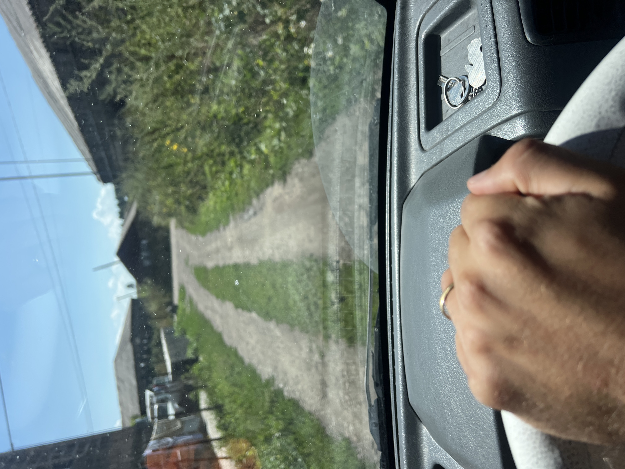 The image is a first-person view from inside a vehicle, looking through the windshield at a dirt road in Khando, Georgia. The road is flanked by green grass and vegetation, with buildings visible on either side. The sky is blue with some clouds. The vehicle's dashboard is visible in the foreground, with a hand resting on the steering wheel and a set of keys in a compartment. The scene appears to be a rural or semi-rural setting.