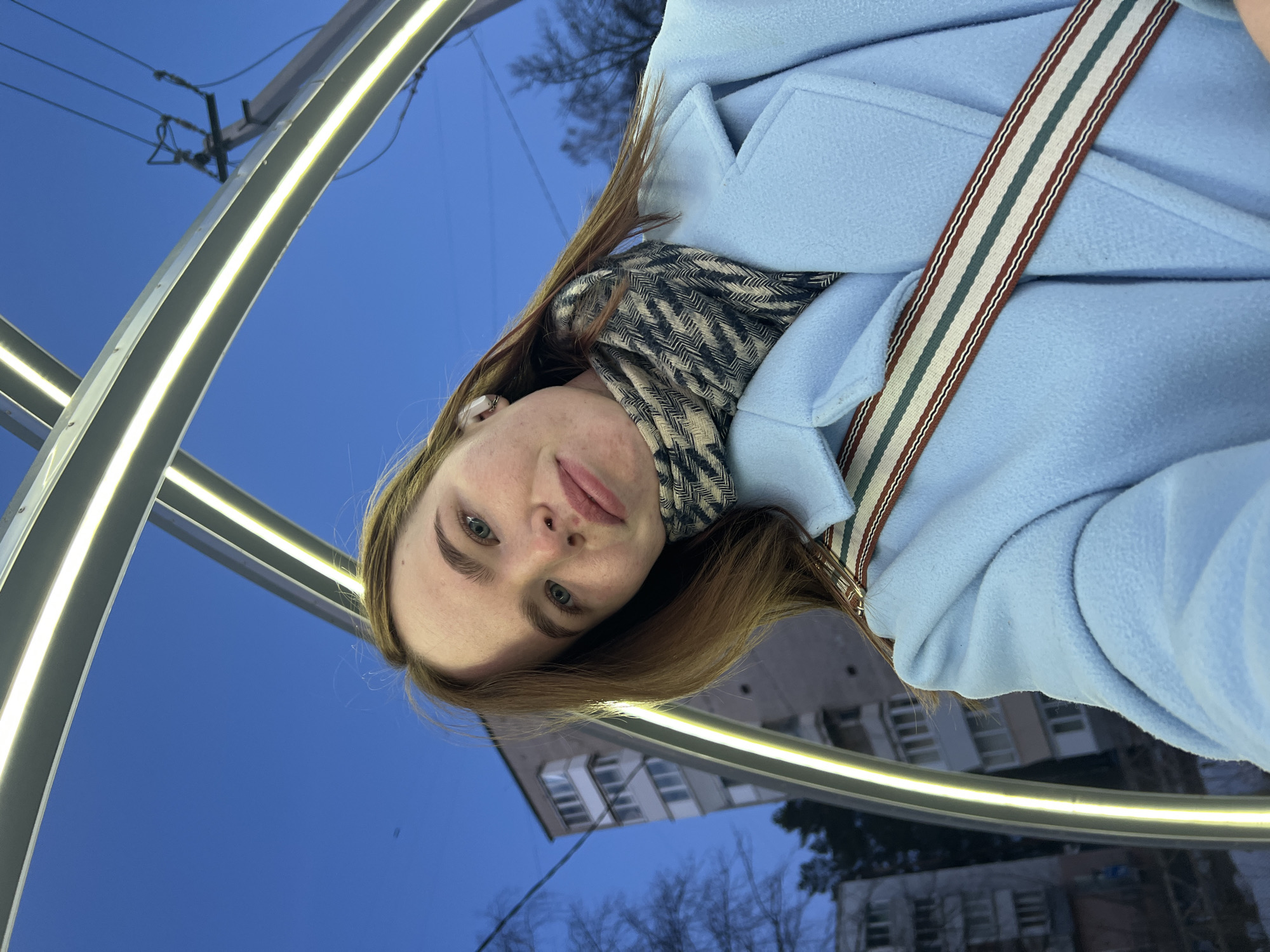 The scene captures a young female subject in a selfie format, standing outdoors at dusk in Chaikovsky, Russia, near the "Light Arch" on Vokzalnaya Street. The female has reddish-brown hair, light blue eyes, visible freckles, and a small stud piercing in her left nostril. She wears a light blue overcoat, a patterned scarf with dark grey and beige herringbone design, and a multi-colored striped strap (white, green, reddish-brown) diagonally across her chest. A white wireless earbud is present in her right ear. She looks directly at the camera with a neutral expression. Overhead, a luminous architectural structure consisting of multiple parallel light strips forms an arch, providing significant foreground illumination. In the background, against a dark blue sky, a multi-story grey concrete residential building with balconies is visible to the left, and a utility pole with power lines is visible to the right. Bare tree branches are scattered throughout the background.