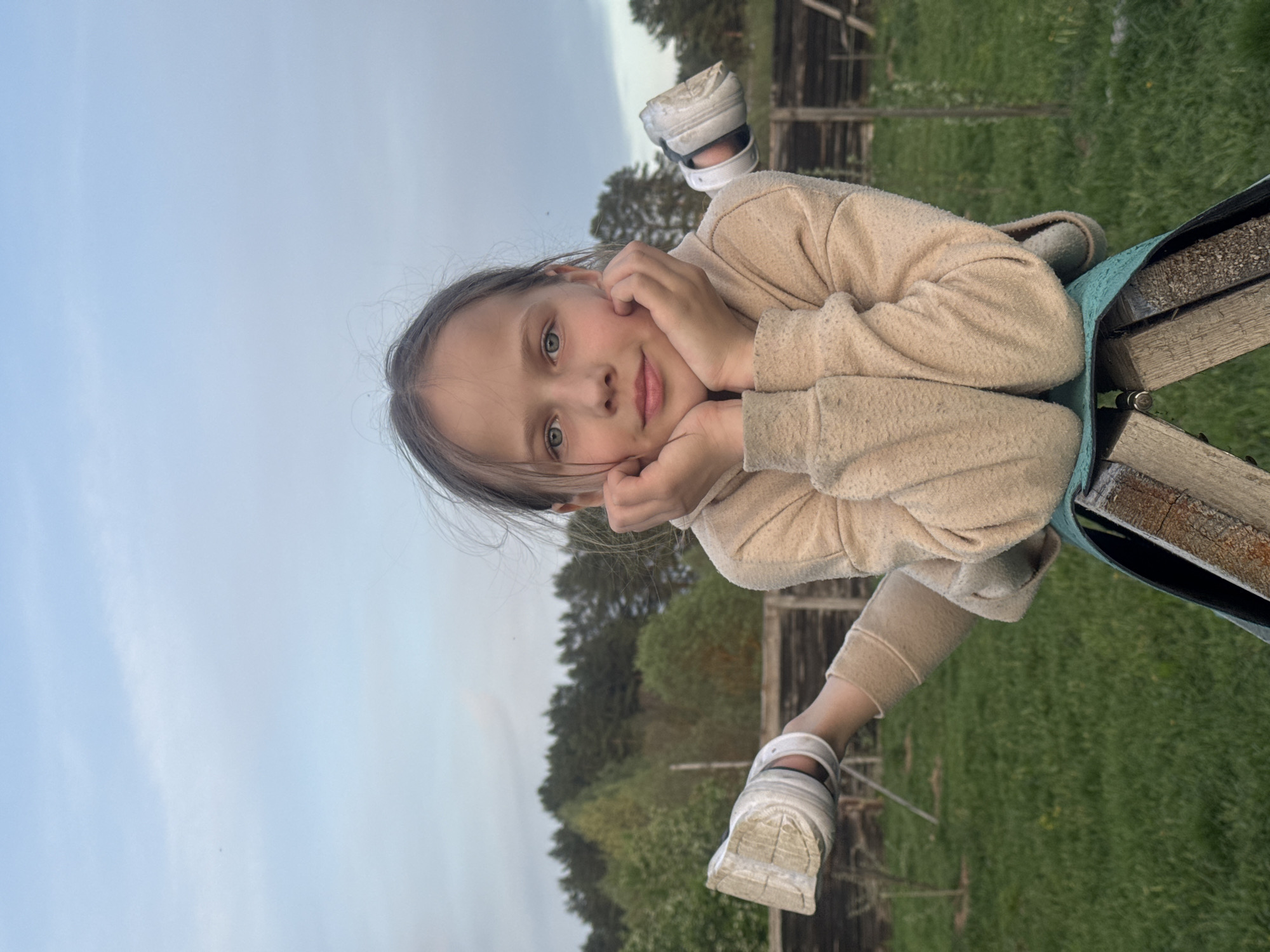 A young girl with light brown hair and blue eyes, wearing a beige sweater and white Crocs, is lying on a swing. She is resting her chin on her hands, looking towards the camera. The swing has a wooden seat and a turquoise support. The background shows a field of green grass, a wooden fence, and trees under a cloudy sky. The specific location in Rusalevka, Russia cannot be confirmed from visual information alone.