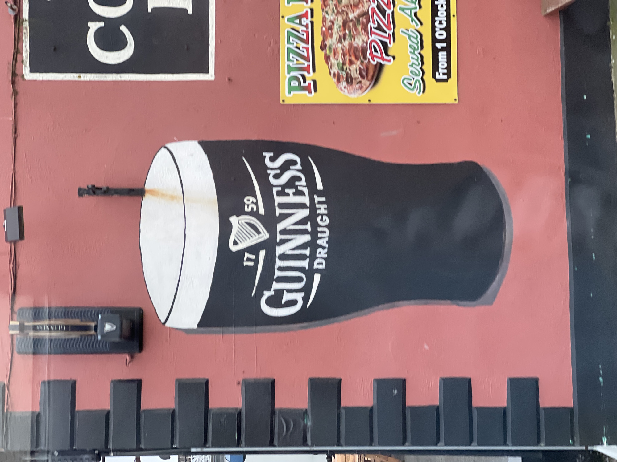The image shows a mural painted on a reddish-brown wall, likely an exterior wall of a building. The mural depicts a large, stylized pint of Guinness beer, with its characteristic black liquid and white head. The Guinness logo is prominently displayed on the glass.
Above the Guinness pint, there are two signs. The one on the left is partially visible and appears to be black with white lettering. The one on the right is yellow with red and green lettering and features an image of a pizza. Text on this sign reads "PIZZA" and "Served At From 1 O'Clock".
Below the Guinness pint, there are black, rectangular, block-like structures that resemble a battlement or a decorative base for the mural. To the left of the Guinness pint, there is a dark object mounted on the wall that appears to be a speaker or a light fixture, with some yellow and black detailing. A thin metal rod extends from the wall towards the Guinness glass.
The overall scene suggests an outdoor setting, possibly the side of a pub or restaurant in Letterkenny, Ireland, given the Guinness branding and the mention of pizza. The lighting and the image quality suggest it was taken during the daytime. No people are visible in the image, and there are no clear indicators of weather conditions. The focus is on the vibrant mural and the signage.