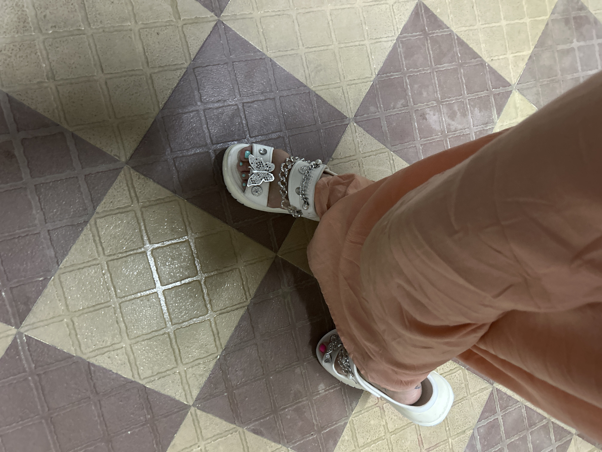 A high-angle shot shows a person wearing peach-colored pants and white sandals adorned with silver butterfly and chain embellishments. The sandals are visible on the right and upper left of the frame. The person is standing on a tiled floor with a geometric pattern of alternating beige and purple square tiles. The scene appears to be indoors. Puttaparthi, India is not discernible from the image.