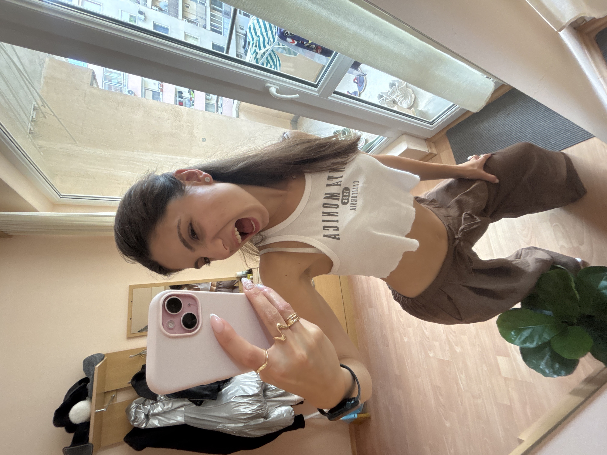 In Varna, Bulgaria, a young woman takes a selfie in what appears to be an apartment. She is wearing a white cropped tank top and brown wide-leg pants. She is holding a pink phone in her right hand, with several rings on her fingers. The background shows a wall with a coat rack holding several jackets, a mirror, and a large window overlooking a city with apartment buildings. A potted plant is visible in the foreground. The woman is smiling with her mouth open.