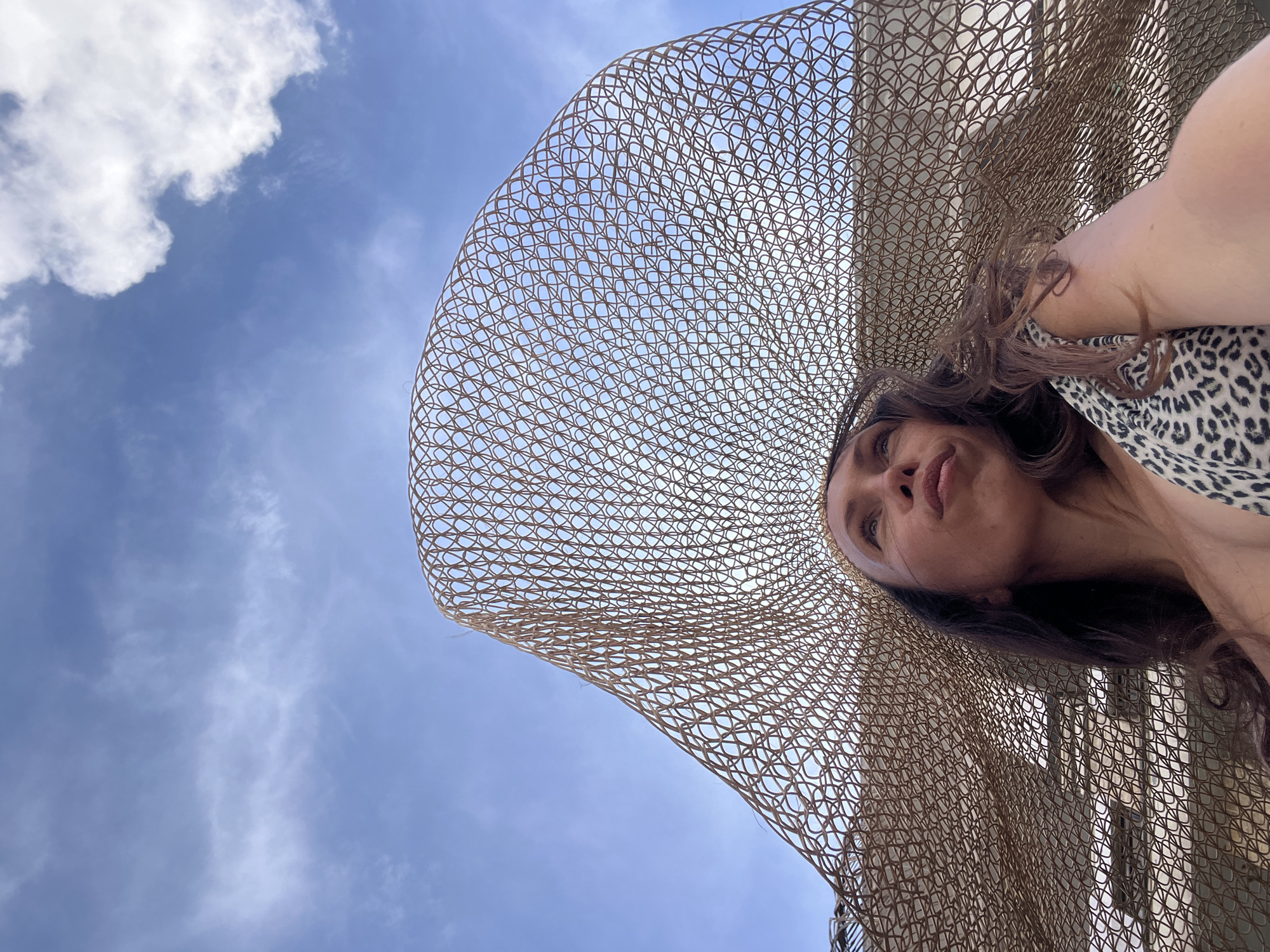 A low-angle shot depicts a woman wearing a large, woven sun hat against a backdrop of a blue sky with scattered clouds. The hat is brown and patterned with circular weaves. The woman is partially visible, with a view of her face, hair, and upper torso, wearing an animal-print top. The location is likely North Kuta, Indonesia.