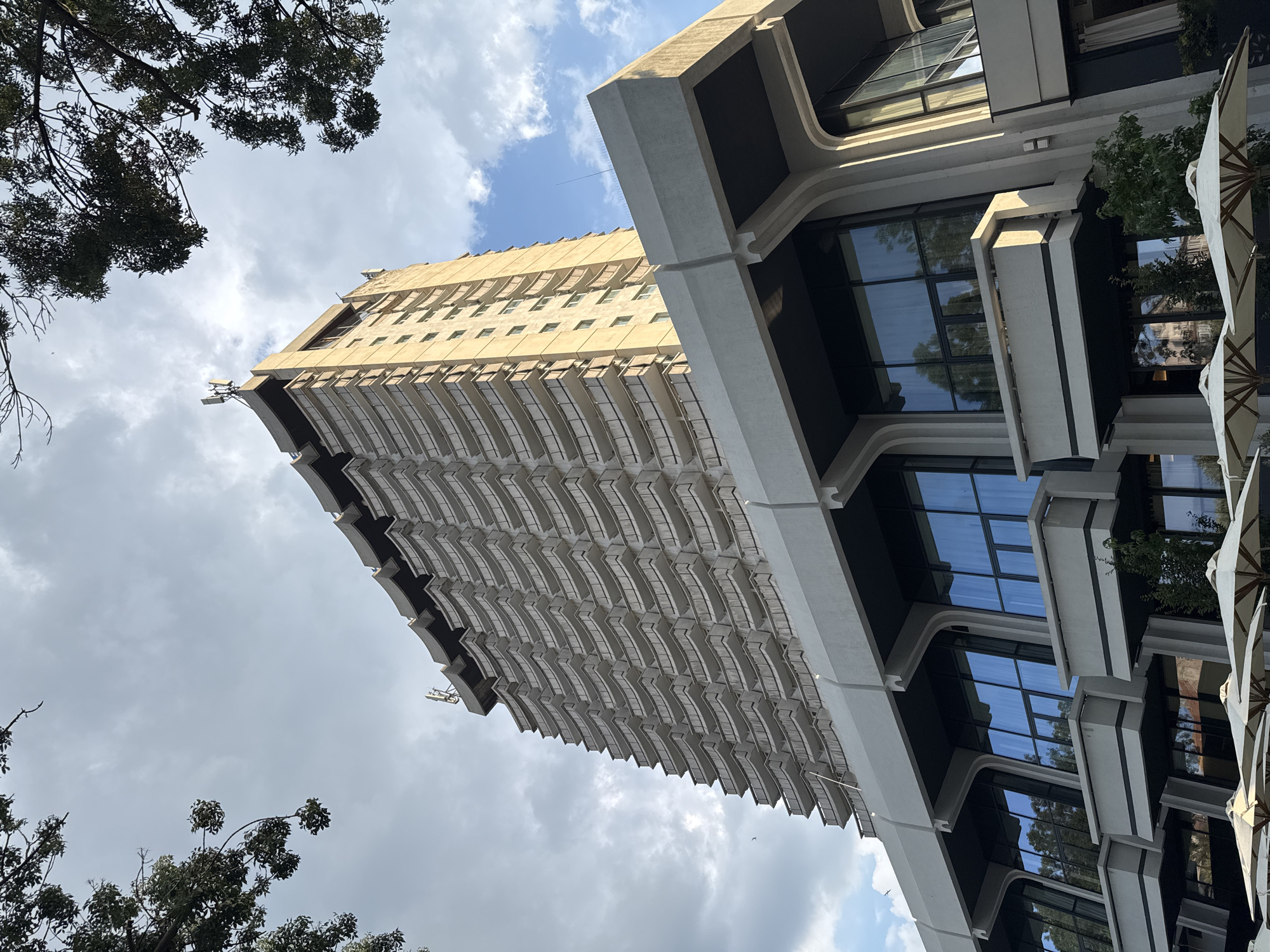 This is a low-angle, full-frame shot of the Grand Hotel Varna, a 1960s Brutalist-style hotel located in the resort complex of Saints Constantine and Helena near Varna, Bulgaria. The hotel is a tall, multi-story structure with a distinctive tiered design, featuring numerous balconies that wrap around the building. The facade is predominantly light-colored, possibly concrete or stucco, with dark-framed windows.
In the foreground, the hotel's lower levels are visible, characterized by large, angular glass windows and projecting concrete structures, some of which appear to be balconies or decorative elements. White umbrellas are arranged in front of these lower levels, suggesting an outdoor seating area. Lush green trees frame the top of the image, with their branches extending into the sky.
The sky is partly cloudy, with patches of blue sky visible amongst the grey clouds. Two small antennas are mounted on the rooftop of the hotel. The overall impression is of a large, imposing hotel structure set against a natural backdrop, with a sense of mid-20th century architectural style. There are no people visible in the image.