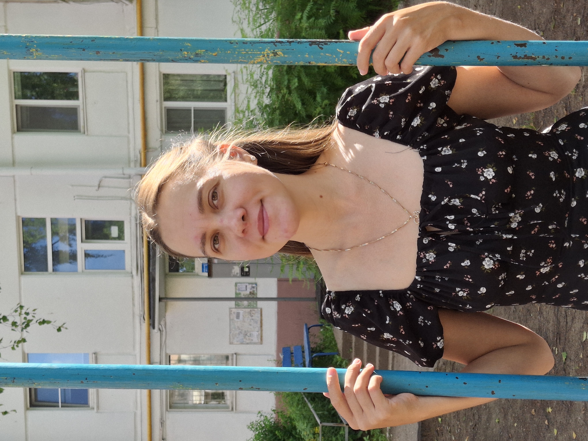 A young woman with long, light brown hair and fair skin is pictured outdoors, leaning against a blue horizontal bar. She is wearing a black dress with a floral pattern of white and pink flowers. Her gaze is directed towards the camera with a slight smile. Her right hand is gripping the blue bar, while her left arm is bent at the elbow and also resting on the bar, with her hand visible. A thin, delicate necklace is visible around her neck.
The background suggests an urban or suburban setting, with a light-colored building with multiple windows visible behind her. Green foliage and trees are present, indicating a natural element within the scene. The blue bar she is leaning on appears to be part of playground equipment or an outdoor exercise structure. The lighting suggests it is daytime, likely sunny, with shadows cast on the ground. There are no visible signs of specific text in the image. The overall impression is one of casual outdoor leisure.