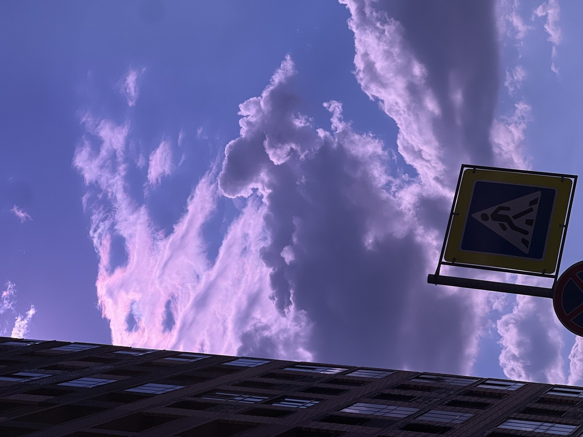 The image is an upward-angled view featuring a building, sky with clouds, and street signs. The building is dark and appears to be multi-storied, with a regular grid of windows. Above the building, the sky is predominantly blue with streaks and patches of clouds, some of which are dark and others are lighter, possibly reflecting sunlight. In the foreground, there are two street signs attached to a metal pole. One is a pedestrian crossing sign (yellow square with a dark silhouette of a person walking), and the other is a no-stopping sign (red circle with a blue background and a white "X"). The scene appears to be captured in Saint Petersburg, Russia. The light suggests either early morning or late evening. There are no visible people or actions taking place.