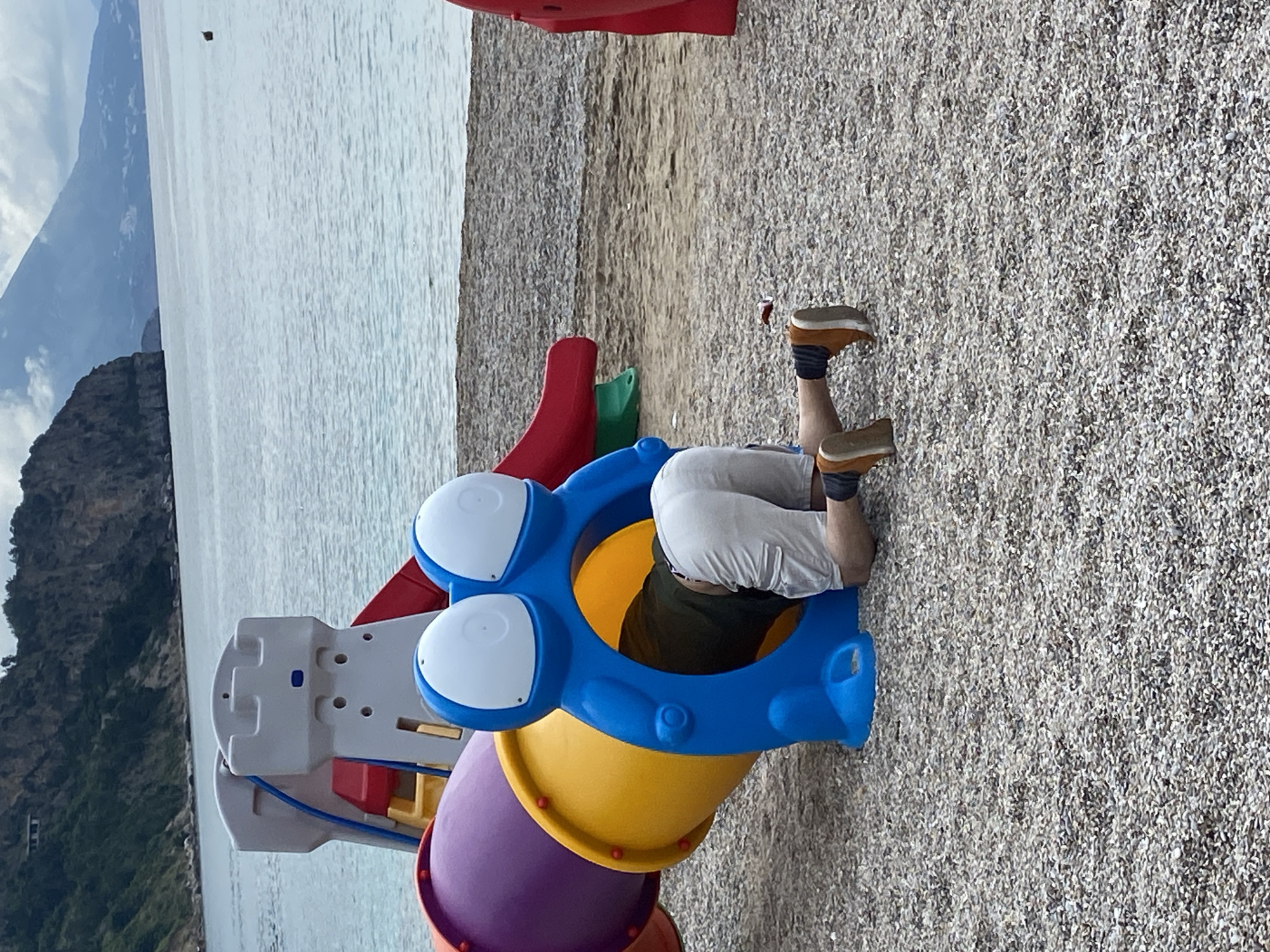 A man is inside a playground structure at a beach in Prijevor, Montenegro. The playground is comprised of a blue, yellow, and purple tube-like slide on the beach. The man's feet are visible, wearing brown shoes and black socks. The beach is made of small pebbles, and in the background, there is water, mountains, and clouds. A small island is visible in the water.