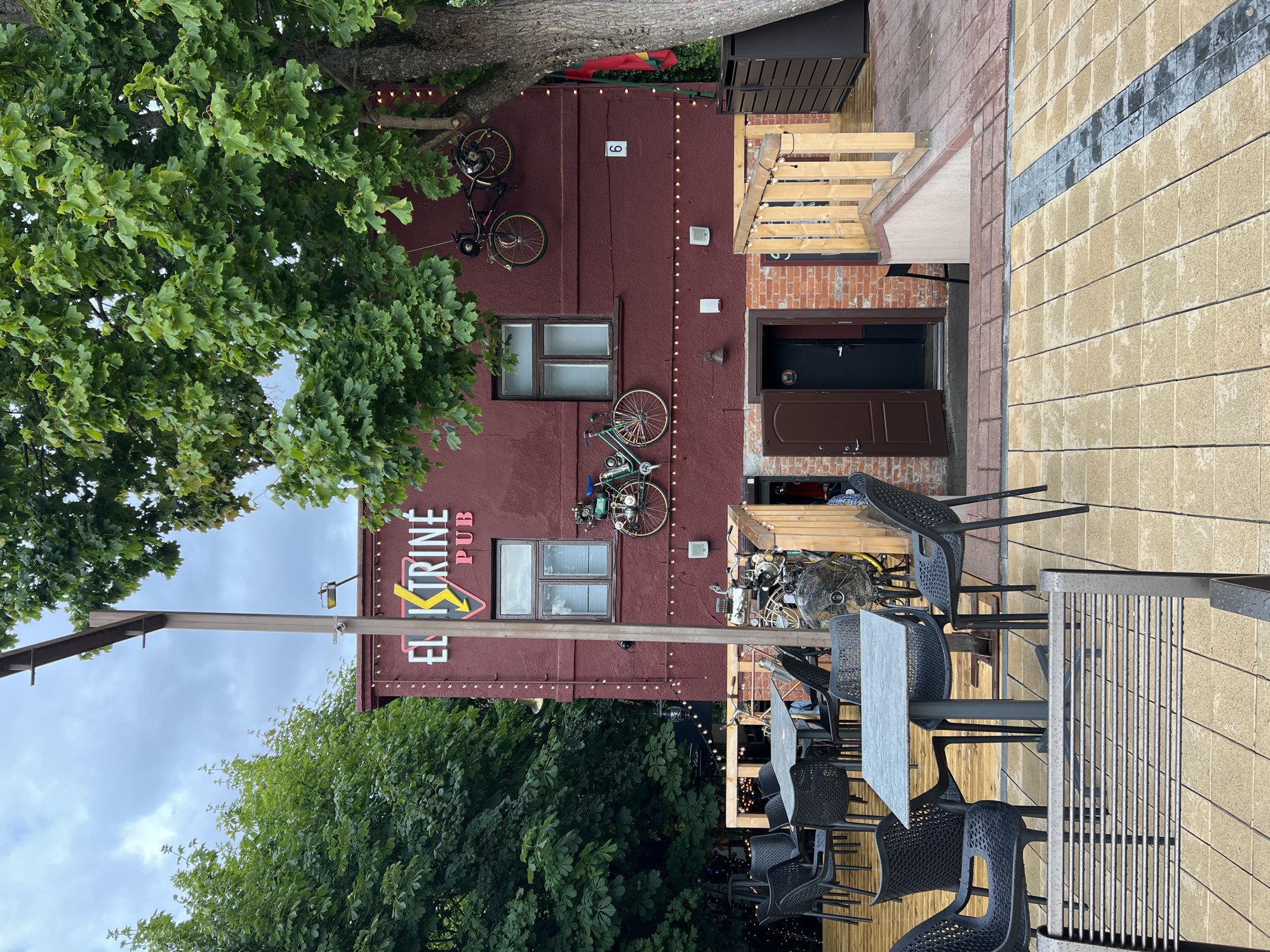 The image shows the exterior of a building with a sign that reads "ELEKTRINĖ PUB". The building is painted dark red and has several windows and doors. There are tables and chairs set up outside, suggesting it is an outdoor seating area for a bar or restaurant. Two bicycles are mounted on the wall of the building as decoration. A Lithuanian flag is visible on the right side of the building. The ground is paved with yellow and grey square tiles. In the background, there are trees and a cloudy sky. The building appears to be located in the city of Palanga, Lithuania.