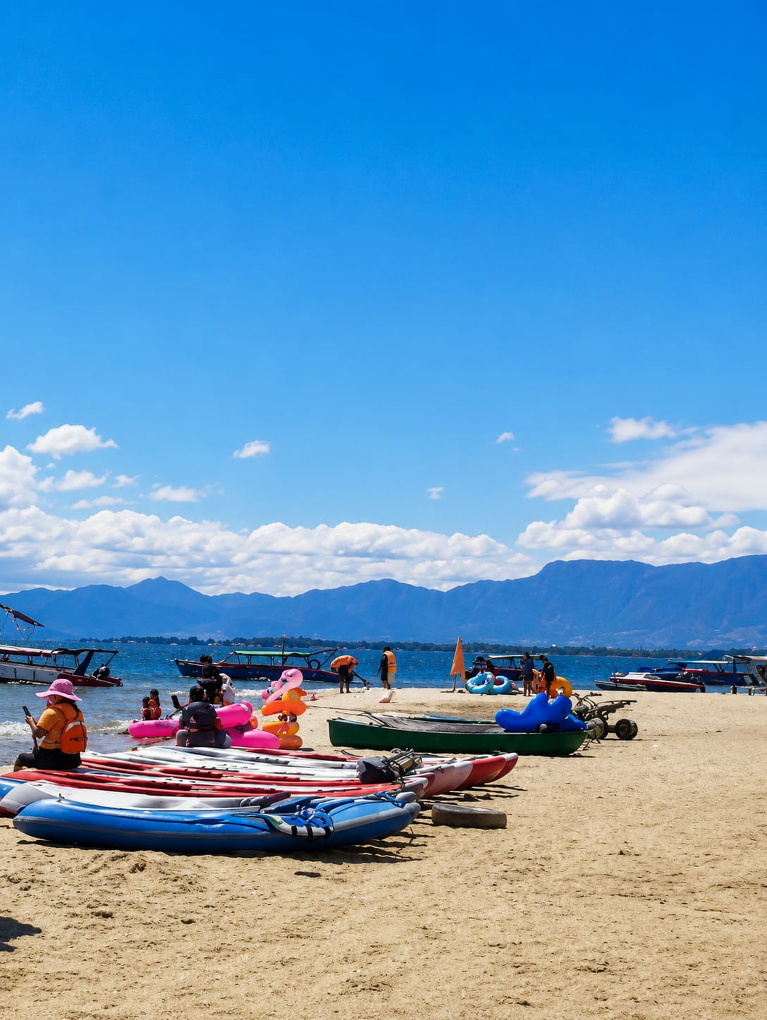 This is a photograph taken at Lumban Bul Bul, Indonesia, a location known for its beach and water activities. The image depicts a sandy beach with several inflatable kayaks and canoes lined up. In the background, the clear blue water of a lake or sea is visible, dotted with various boats. Beyond the water, a mountain range under a bright blue sky with scattered white clouds forms the horizon.
On the beach, there are several people engaged in leisure activities. A woman wearing a pink hat and an orange vest is seated on the sand, looking at her phone. Other individuals, some wearing orange life vests, are scattered around, with some interacting with inflatable toys in the shallow water. A few people are standing near the water's edge, and others are seated on the sand.
The overall atmosphere is one of a sunny day at a recreational beach, with various watercraft available for rent or use. The presence of mountains in the distance suggests a scenic natural environment.