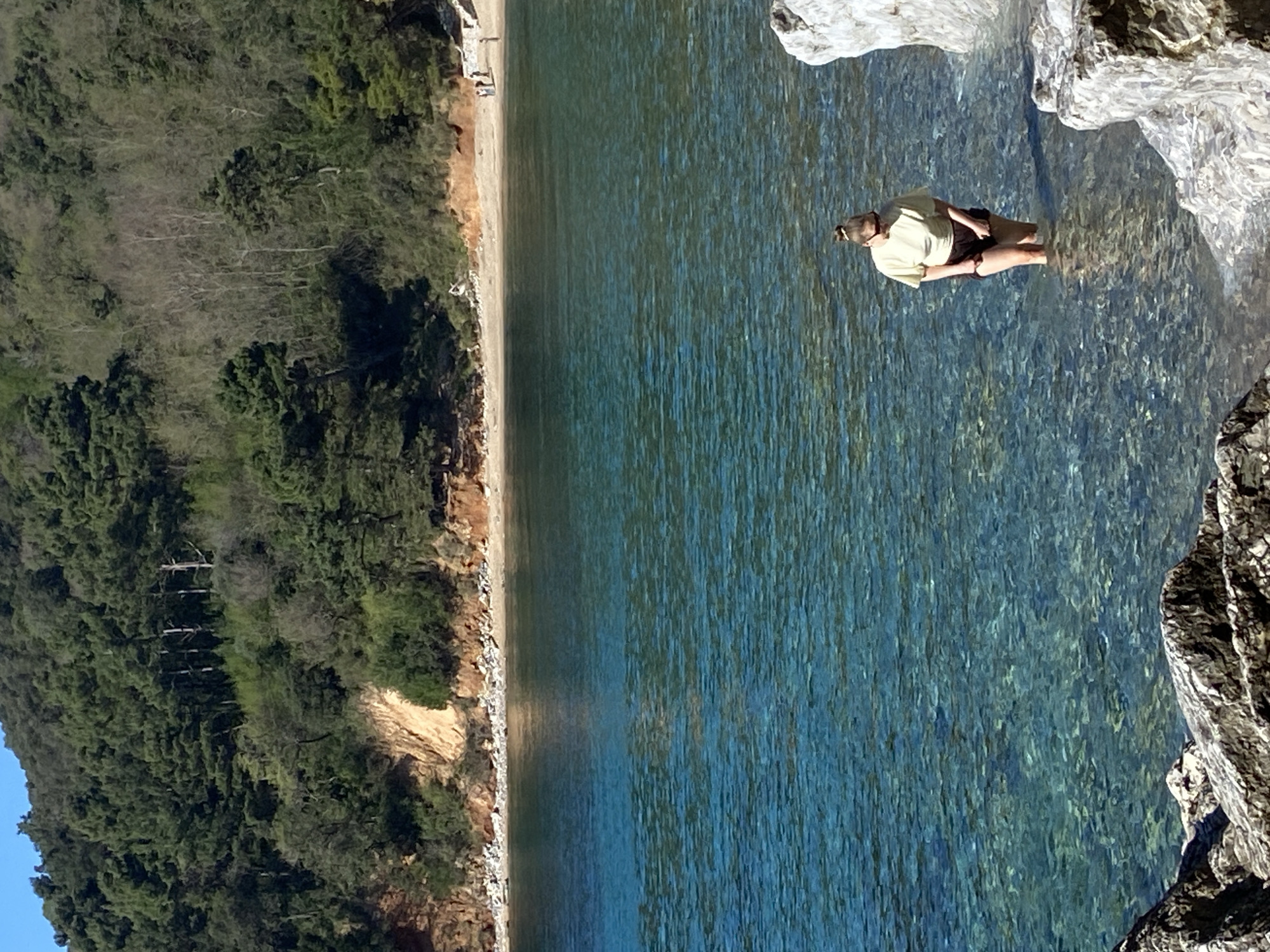 The image depicts a woman standing in clear, shallow water near a rocky coastline. She is wearing a light yellow short-sleeved shirt and dark shorts. Her hair is pulled back in a bun. The water is clear enough to see the bottom, and the surrounding area features a sandy beach bordered by a forest of green trees. The location is likely near the city of Budva, Montenegro, given the coastal terrain and vegetation.
