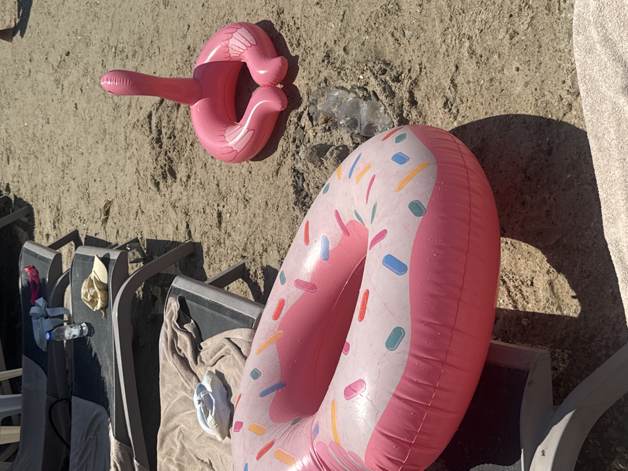 The image depicts a beach scene in Bodrum, Turkey, with a pink inflatable donut pool float prominently placed on the sand. Adjacent to the donut float is a pink inflatable flamingo. In the background, two empty lounge chairs are visible, one with a towel, a water bottle, and a tan hat on it. The sand is light brown and appears dry, with some small pebbles and debris scattered around. The lighting suggests a sunny day.