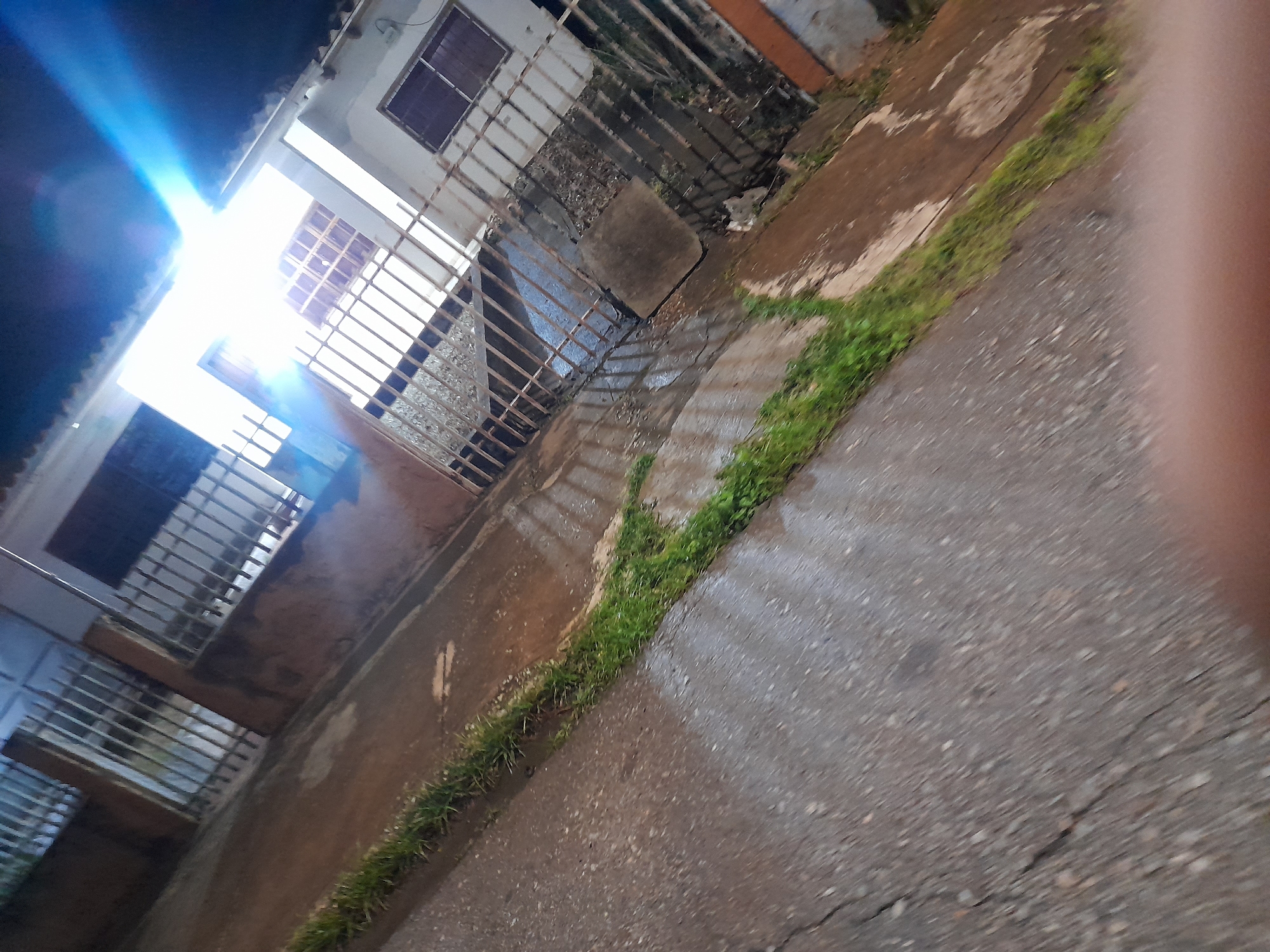 The image shows a street scene at night in Santa Ana, Venezuela. The perspective is low and angled, with a bright light source dominating the upper right quadrant, causing lens flare. The foreground is a cracked, wet asphalt road. To the left and extending towards the background is a patch of green grass growing along the edge of the road. Beyond the grass is a brown, dirt-like area. In the background, there is a building with a white facade and barred windows and gates. The overall impression is one of a poorly lit, somewhat neglected urban environment.