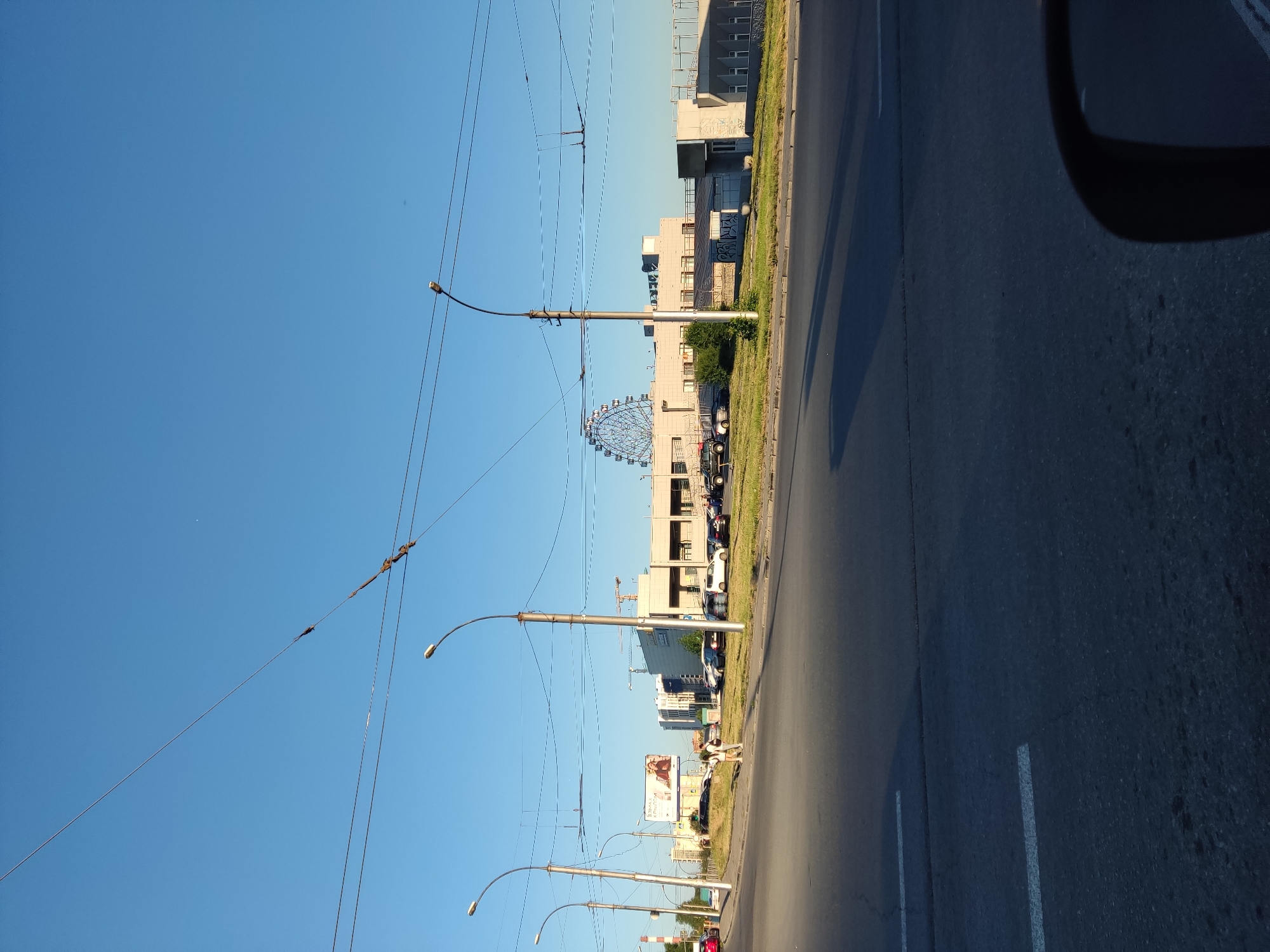 This media file captures a street scene in Novosibirsk, Russia. The image is oriented vertically, with the sky occupying the left side and the road and buildings on the right. A large Ferris wheel is visible in the distance, a prominent landmark against the clear blue sky. Several tall streetlights with overhead wires run parallel to the road, suggesting an urban or suburban environment with public transportation like trams or trolleybuses.
Several buildings, varying in height and architectural style, line the street. Some appear to be modern commercial structures, while others have a more industrial or residential look. There are cars parked along the roadside and a few people are visible walking on the sidewalk. One person is seen standing near a billboard. The time of day appears to be late afternoon or early evening, judging by the long shadows cast across the road and the warm sunlight. The weather seems clear and pleasant.
There is some visible text on a billboard in the middle ground, but it is not legible enough to be identified. The overall impression is a typical day in a Russian city, with a mix of modern infrastructure and everyday life unfolding. The presence of the Ferris wheel suggests it might be a recreational area or a part of a larger entertainment complex.