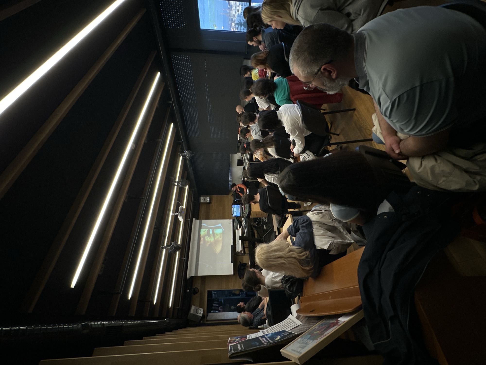 The image depicts an indoor scene within a modern tiered auditorium, lecture hall, or cinema, likely in Sofia, Bulgaria. Numerous individuals are seated, primarily viewed from behind or the side, facing forward towards a large projection screen at the front of the room. The screen displays an image or video, possibly featuring a human figure or face within a natural setting. Above the main screen, another smaller display or window shows an outdoor, possibly urban, scene.
The architecture of the hall features dark walls and ceiling, accented by bright, linear LED light strips that run diagonally, creating a distinctive modern aesthetic. Wooden elements are visible in the seating and wall paneling. Mounted speakers and projectors are also part of the technical setup.
In the immediate foreground, to the right, there are stacks of books, magazines, or brochures on a wooden surface. One visible publication cover appears to have the word "EXPLORERS" or "EXPLORE" partially legible. A man with a beard and glasses, wearing a light blue polo shirt, is seated in the foreground, his attention directed forward and slightly downwards, mirroring the focus of the other attendees.
The overall atmosphere suggests an attentive audience engaged in watching a presentation, lecture, or film screening. The lighting is controlled, likely dimmed to enhance the visibility of the projected content.