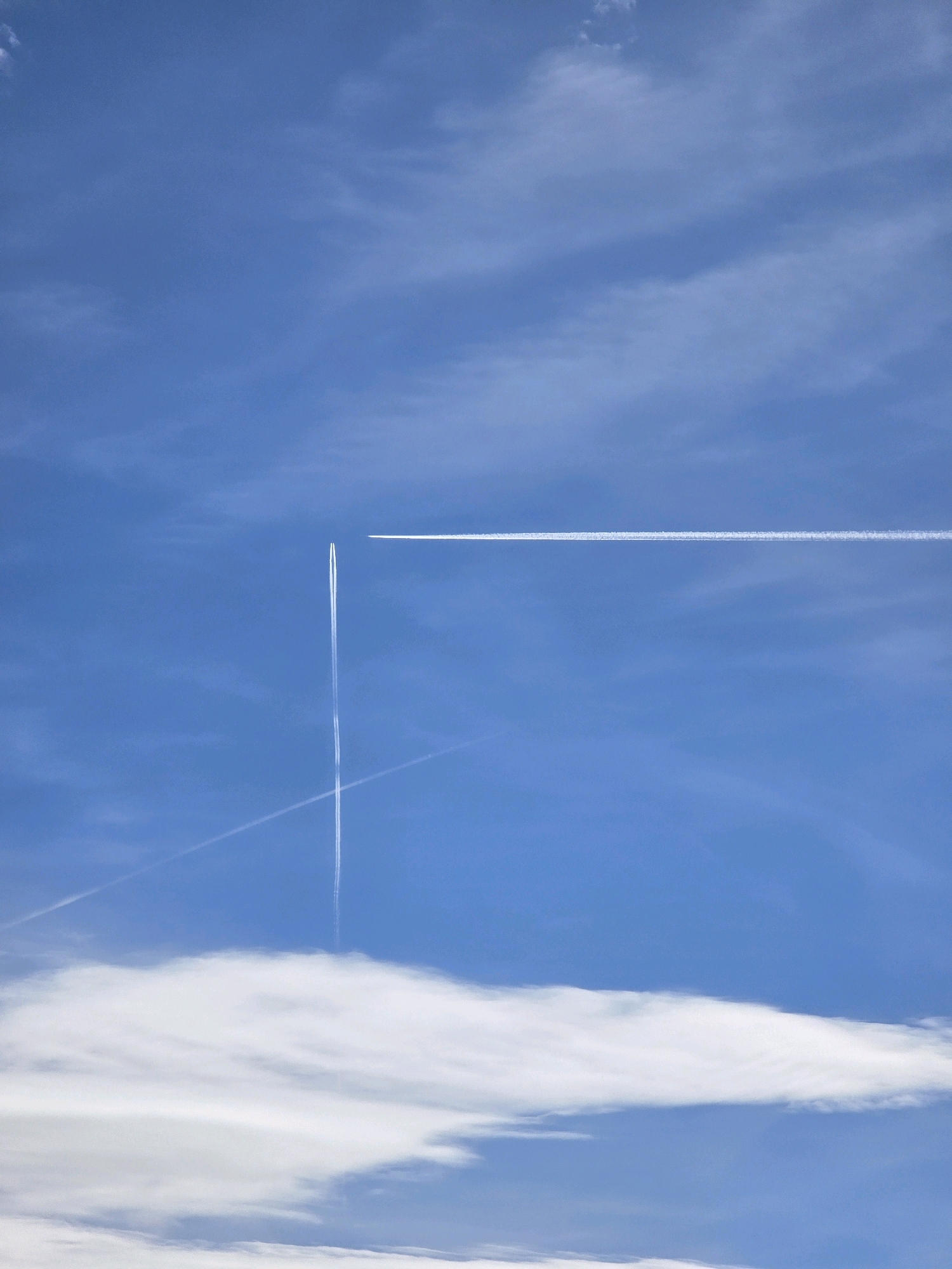 The image depicts a clear blue sky above the city of Garmisch-Partenkirchen, Germany. Two distinct contrails from airplanes are visible, crossing each other in the sky. One contrail stretches horizontally across the upper right portion of the frame, while the other forms a vertical line with a diagonal extension in the lower left quadrant. A large, fluffy white cloud formation occupies the bottom of the image. There are no people or buildings visible in this aerial view.