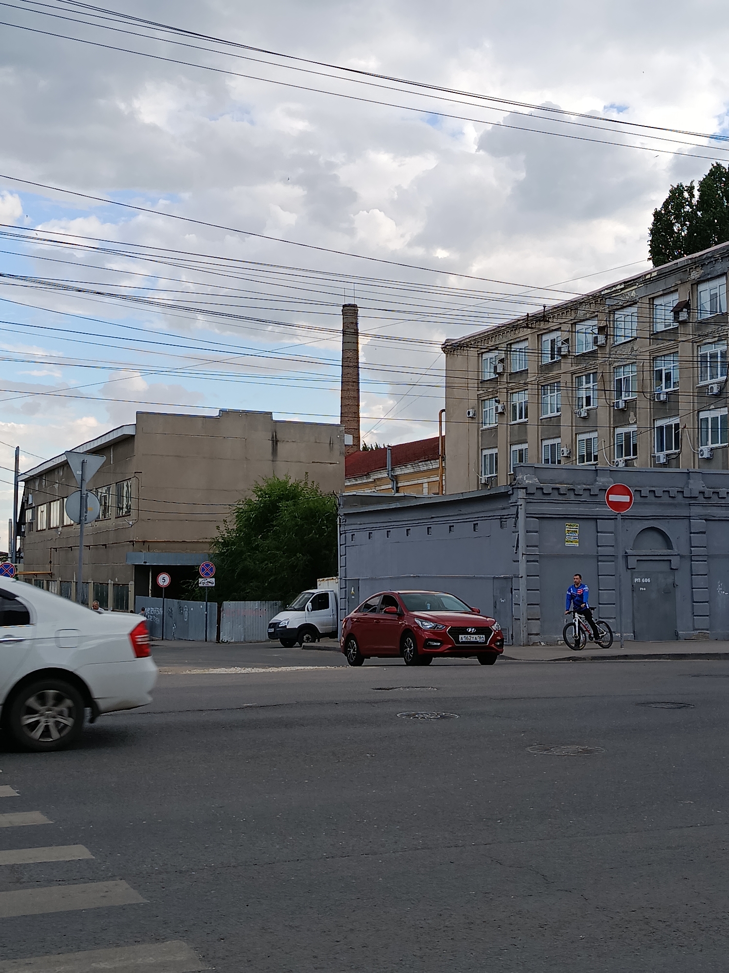 This is a street-level, eye-level, medium shot of a street scene in Saratov, Russia, on a cloudy day. In the foreground, a white car is partially visible on the left side of the frame, moving away from the camera and crossing a pedestrian crossing. To the right of the white car, a red sedan is stopped at an intersection. The sedan has a license plate that reads "A162TA 164". Further to the right, a person is riding a bicycle.
In the background, there are several buildings. On the left, a long, beige industrial-style building with multiple windows is visible. To its right, a tall, red brick smokestack rises into the sky. In the middle ground, a grey, two-story building with a patterned facade and arched doorways stands. Above this building, a larger, multi-story building with numerous windows and visible air conditioning units stretches across the frame. Overhead, a complex network of power lines is strung across the sky.
Several traffic signs are visible, including a red and white circular "no entry" sign on the grey building. Other signs are present on poles near the beige building. The overall environment appears to be an urban industrial or commercial area.