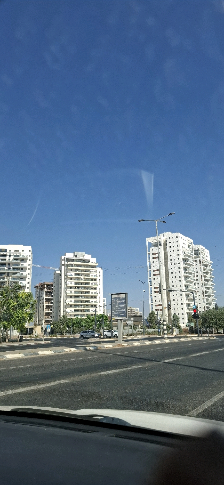 The image captures a street scene in Be'er-Sheva, Israel, with a clear blue sky and a few wispy clouds. The foreground shows a road with a lane marking and a portion of a car's dashboard and windshield wiper. Beyond the road, a sidewalk with a curb separates it from a strip of green trees and grass.
Dominating the background are several modern, multi-story white apartment buildings, some of which appear to be under construction, indicated by a crane visible behind one building. A prominent street sign with Hebrew text stands near the center of the frame. Traffic lights and street lamps are also visible. Two cars are parked on the side of the road near the sign, and another car is driving on the street. The architecture of the buildings, particularly the balconies, suggests a contemporary residential area in Israel. The overall impression is one of urban development under a bright, sunny sky.