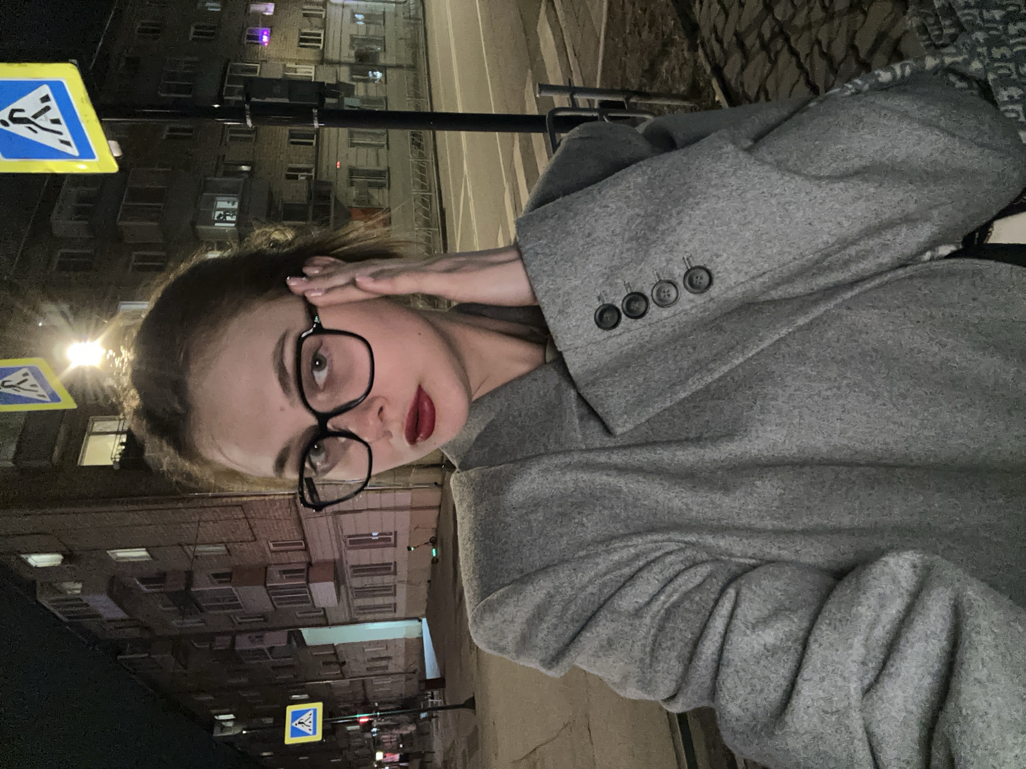 A woman with glasses and dark lipstick poses in a gray coat on a street in Krasnoyarsk, Russia. The background shows buildings, streetlights, and pedestrian crossing signs.