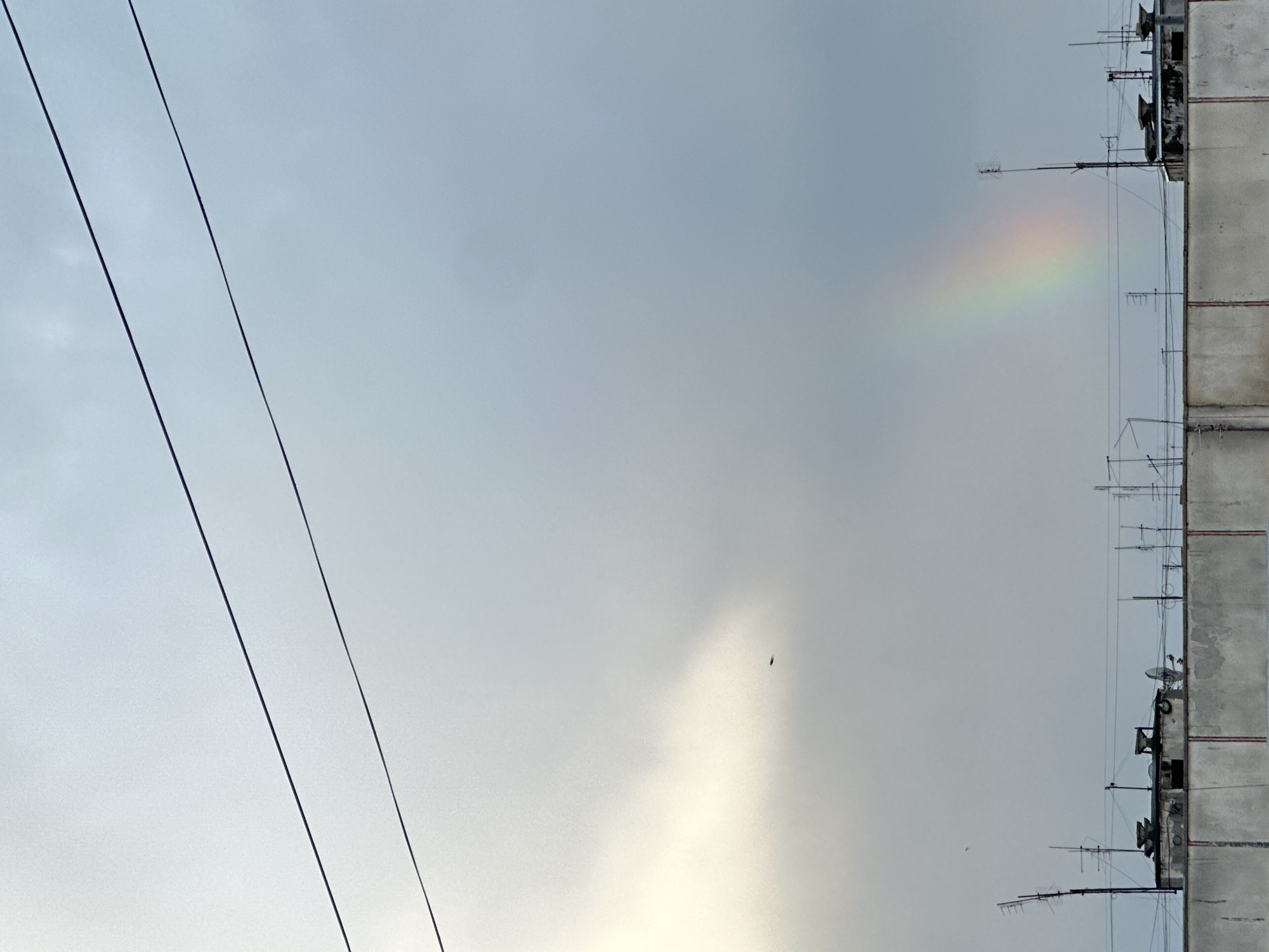 The image depicts a section of an urban environment, likely in a city setting within the Unknown region. The scene is dominated by an overcast sky, with a visible rainbow segment positioned somewhat centrally. In the lower center of the image, a small dark shape, possibly a bird, is visible against a brighter patch of sky. Along the left side, two power lines run diagonally from the upper left towards the lower left. On the right edge, the side of a concrete building is visible, featuring multiple antennas and satellite dishes affixed to the structure.