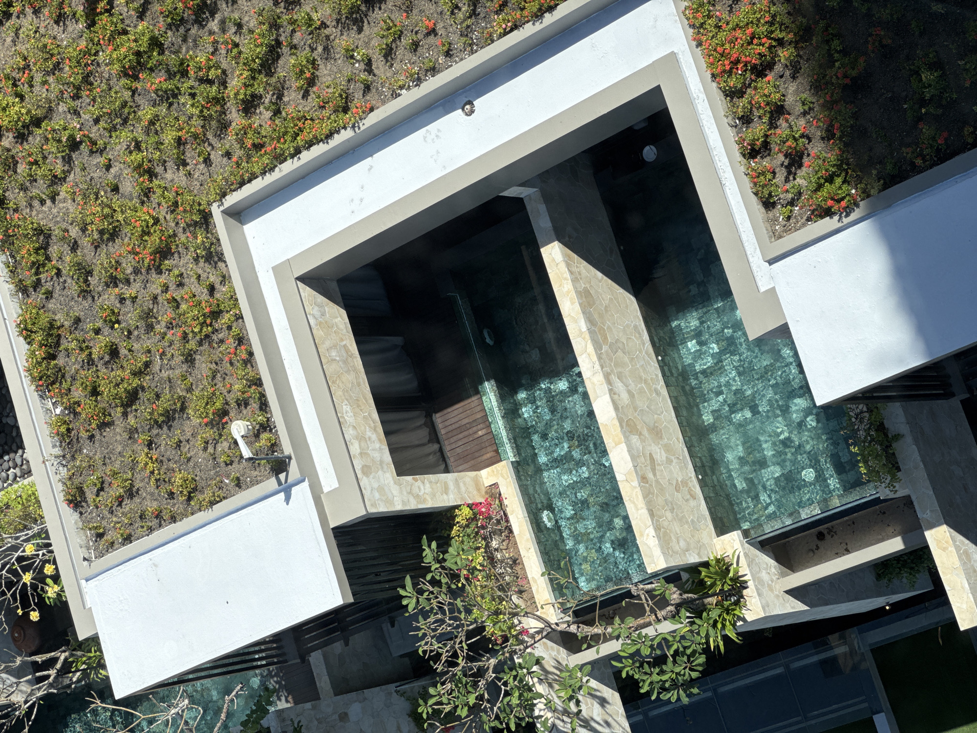 An overhead view depicts a building with two pools situated in Kuta Selatan, Indonesia. The structure is predominantly white with light tan stone accents. Each pool is sectioned by a stone wall, creating distinct zones within the water. One section of the roof is covered in low-growing vegetation with small red flowers, giving it the appearance of a green roof. Trees and other plants are visible around the building.