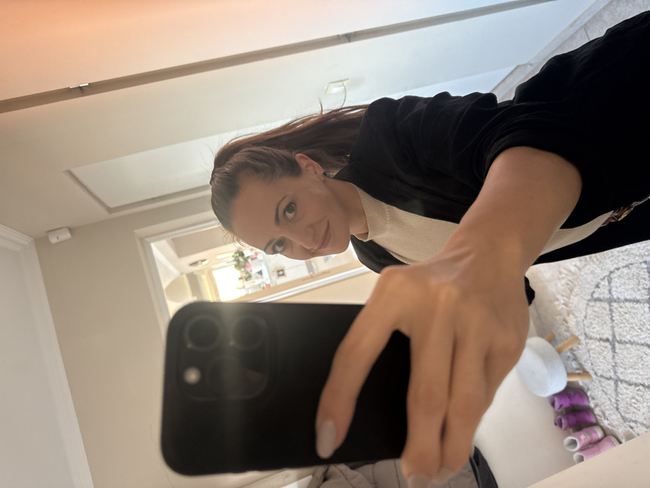 A female subject is depicted taking a mirror selfie indoors, holding a black smartphone with a multi-lens camera array. The woman has brown hair tied in a high ponytail, a light complexion, and is wearing a light cream-colored ribbed turtleneck shirt under a dark blazer. Her visible fingernails are painted a light, neutral color. She is looking towards the camera with a slight smile.
The reflected environment is a residential hallway or entryway. The walls are light beige, and the ceiling is white with decorative molding. A white security sensor is mounted on the upper portion of a wall. Two doorways are visible in the background; one leads to a room with a white door, and another opens to a brightly lit area showing a window and a white console table with decorative elements, possibly flowers. The floor is covered by a light-colored rug with a geometric pattern. On the rug, a small, light-colored stool with wooden legs is present, alongside several pairs of children's shoes, predominantly pink and purple, neatly arranged. The image contains no visual information to determine a specific location within the city of Varna, Bulgaria.