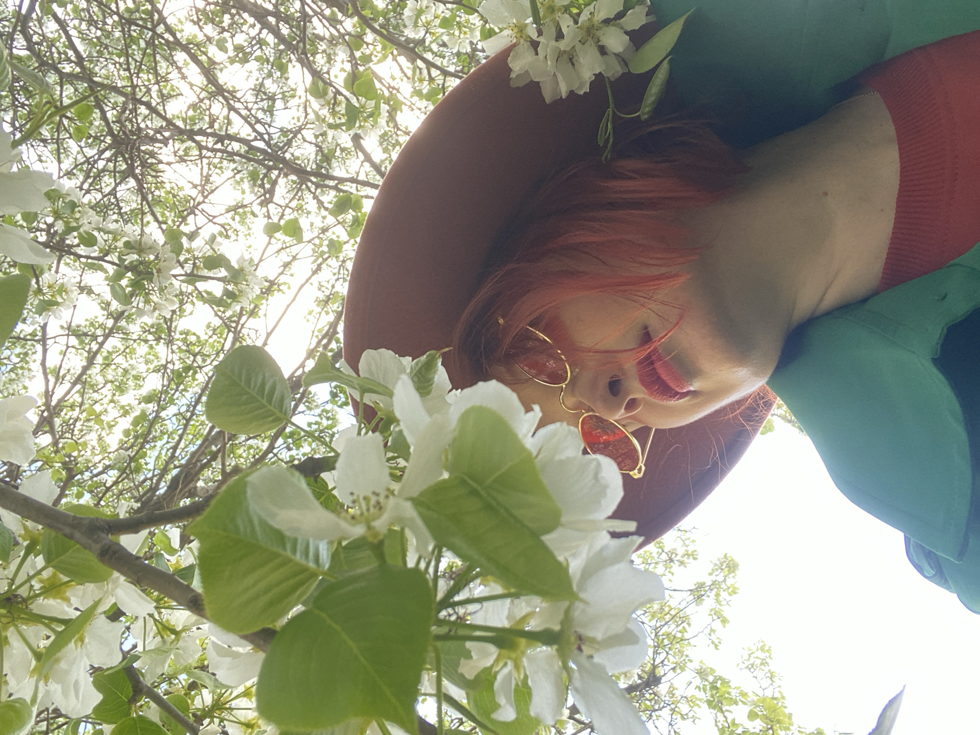 A low-angle shot features a woman with red hair, wearing a brown hat and red-tinted sunglasses. She is also wearing a red sweater under a green jacket. The photo is taken upwards, looking at her face against a backdrop of white blooming trees, likely in a park or garden. The specific location in Yekaterinburg is not identifiable from the image.