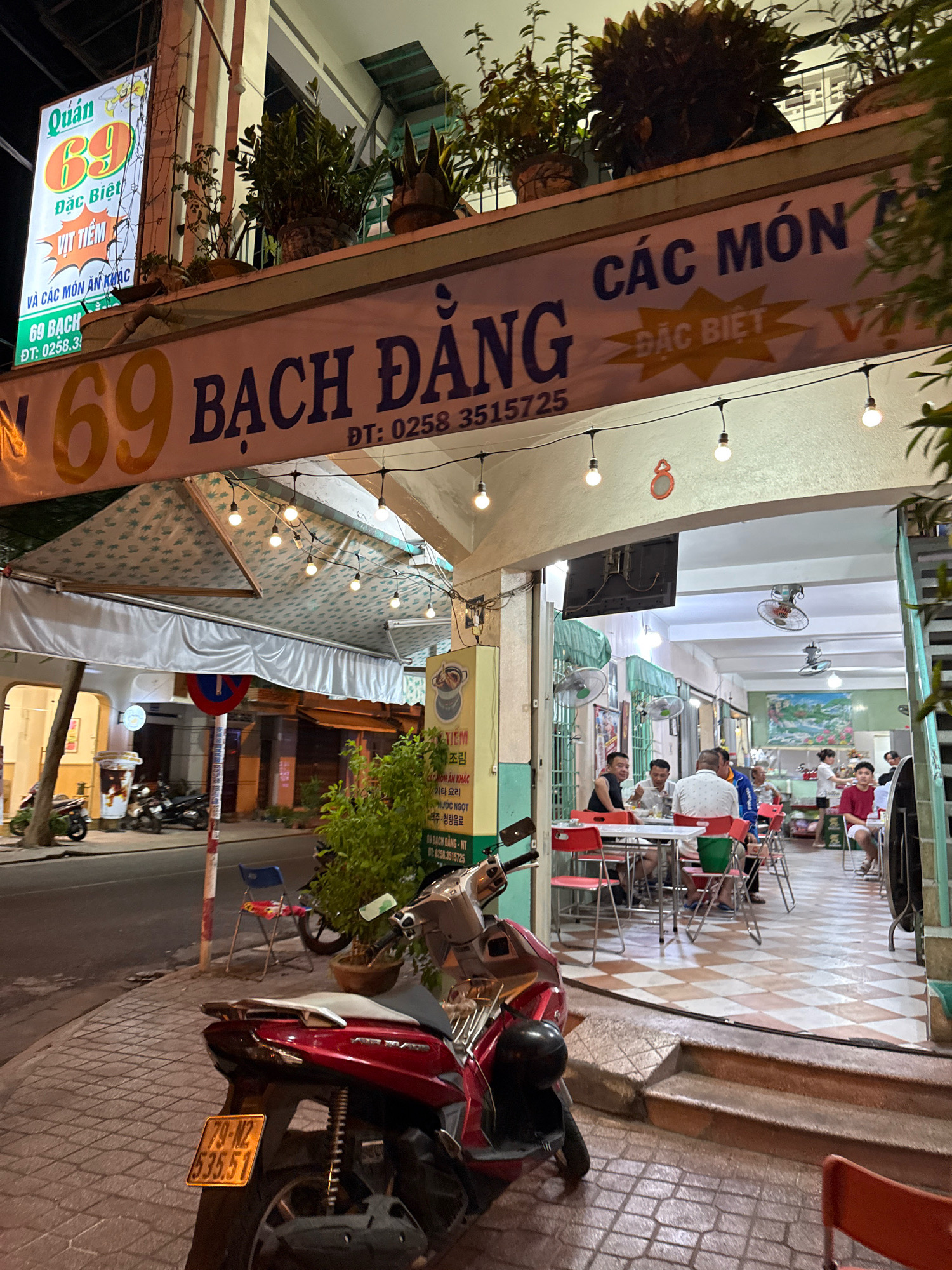 This image depicts a street-level view of a Vietnamese restaurant at night, likely in Nha Trang, Vietnam, given the signage. The restaurant, identified by the prominent "69" logo, features an outdoor seating area with red chairs and tables. A large banner above the entrance reads "QUÁN 69 ĐẶC BIỆT VỊ TIỀM VÀ CÁC MÓN ĂN KHÁC 69 BACH ĐANG ĐT: 0258.3515725". Inside the restaurant, several patrons are seated at tables, some eating, and a few staff members are visible. The scene is illuminated by overhead lights and string lights. A red scooter with license plate "79-N2 535.51" is parked on the sidewalk in the foreground. The building facade shows architectural elements typical of Vietnamese street-front establishments.