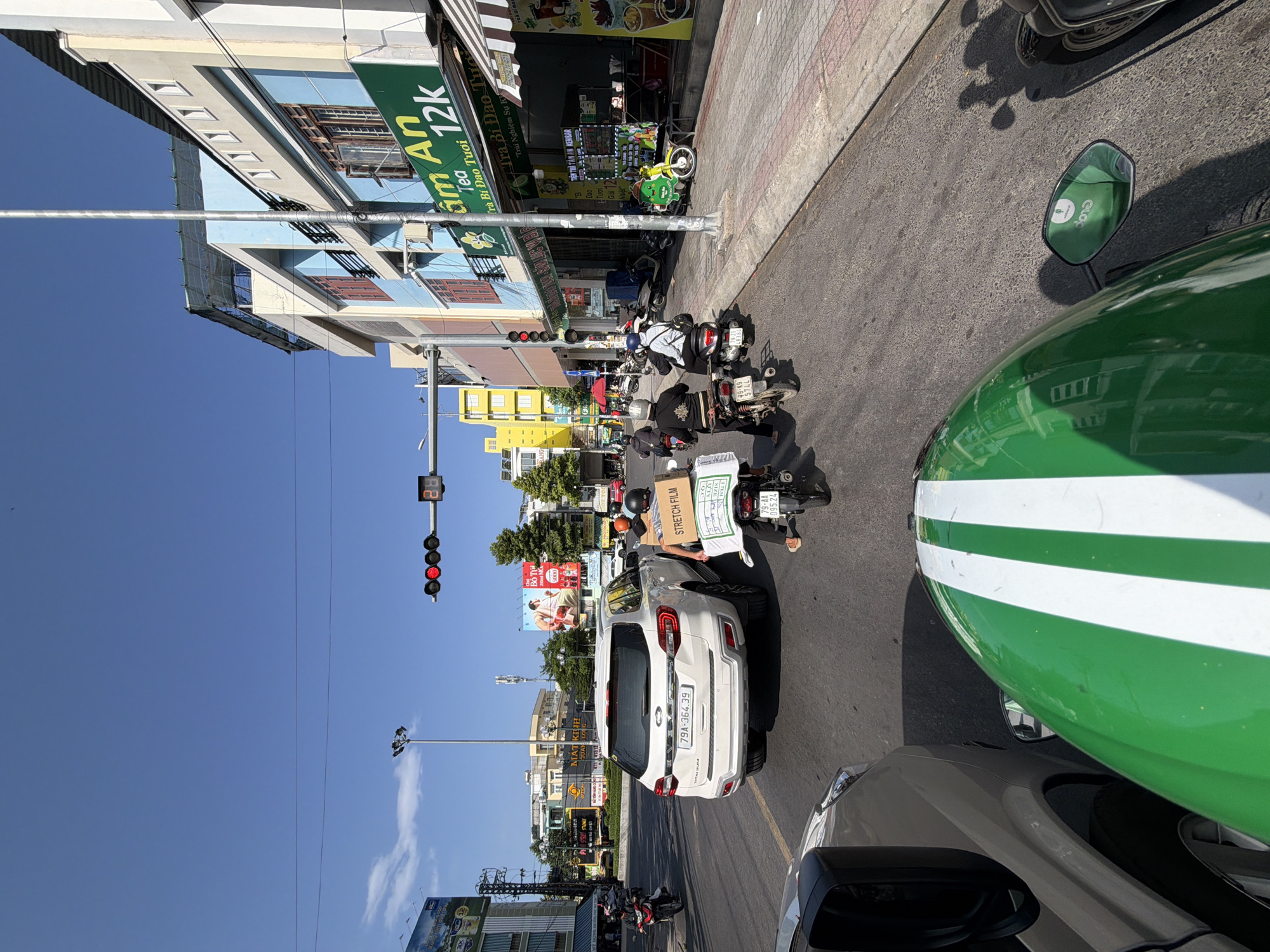 The image captures a street scene in Nha Trang, Vietnam, on a clear, sunny day. The perspective is from a motorbike, looking forward at traffic and the surrounding urban environment. A white SUV, license plate 79A-364.39, is stopped in the foreground. Two individuals on a motorbike are behind the SUV, with one carrying a large cardboard box labeled "STRETCH FILM". Several other motorbikes and their riders are visible in the traffic, moving in the same direction. On the right side of the street, a building with a green awning and signage advertising "Âm An Tea 12k" is prominent. Across the street, another building with a large billboard featuring an advertisement is visible. Traffic lights show a red signal. The road surface is asphalt, and the sidewalk is paved with bricks. Power lines and streetlights are present throughout the scene.