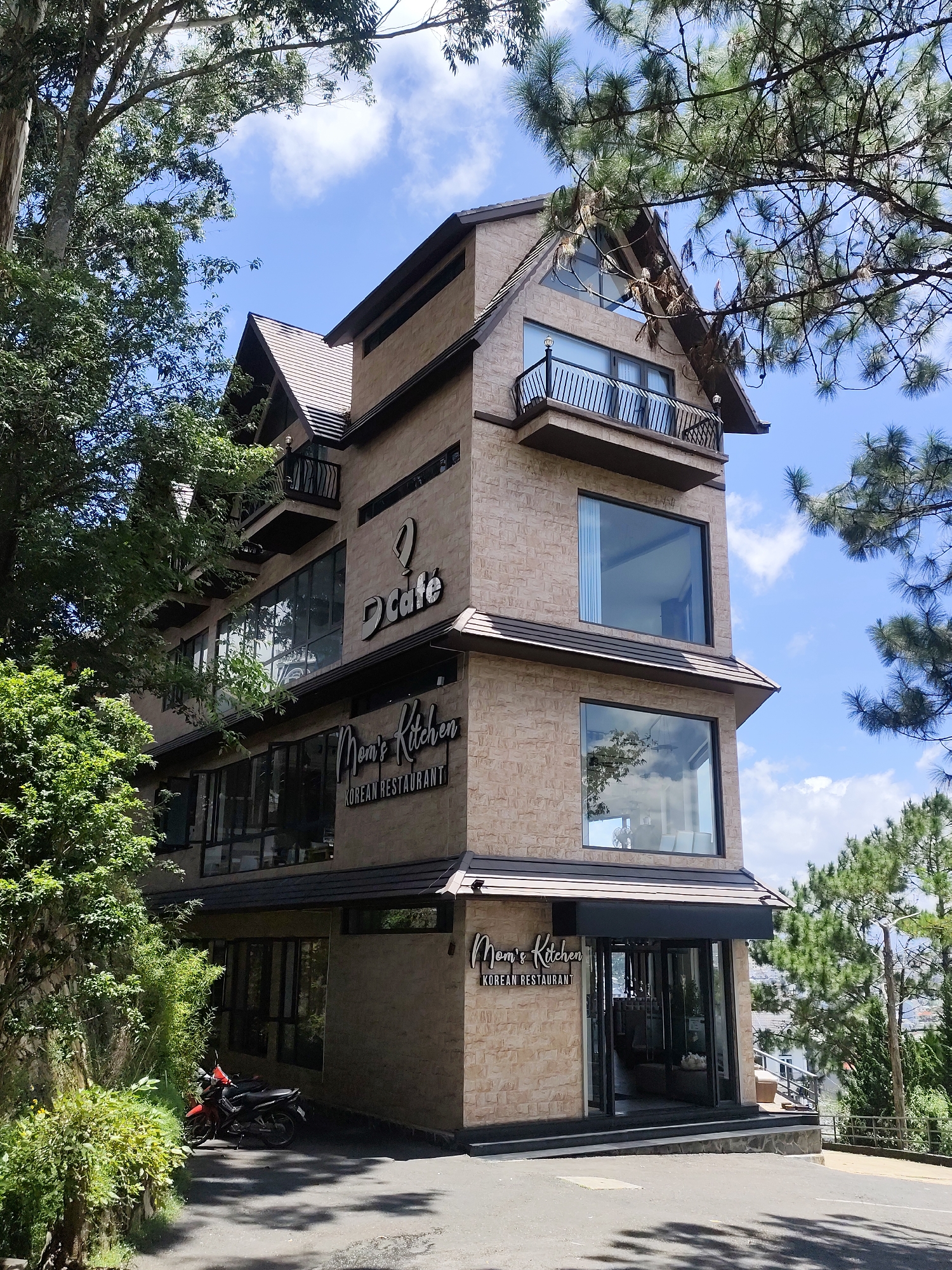 This is a photograph of a three-story building with a modern architectural style, situated on a hillside in Da Lat, Vietnam. The building's exterior is clad in light brown textured stone. It features several large windows, including some with black frames and one with a black balcony railing. The building has a multi-pitched roof with dark brown trim. Signage on the building indicates that it houses "D Cafe" and "Mom's Kitchen Korean Restaurant." The restaurant entrance is at ground level, with double glass doors. The building is surrounded by lush green trees and vegetation, with a blue sky and scattered white clouds overhead. In front of the building, on the paved area, are two parked motorcycles. The overall impression is a serene, possibly slightly elevated, location within the city.