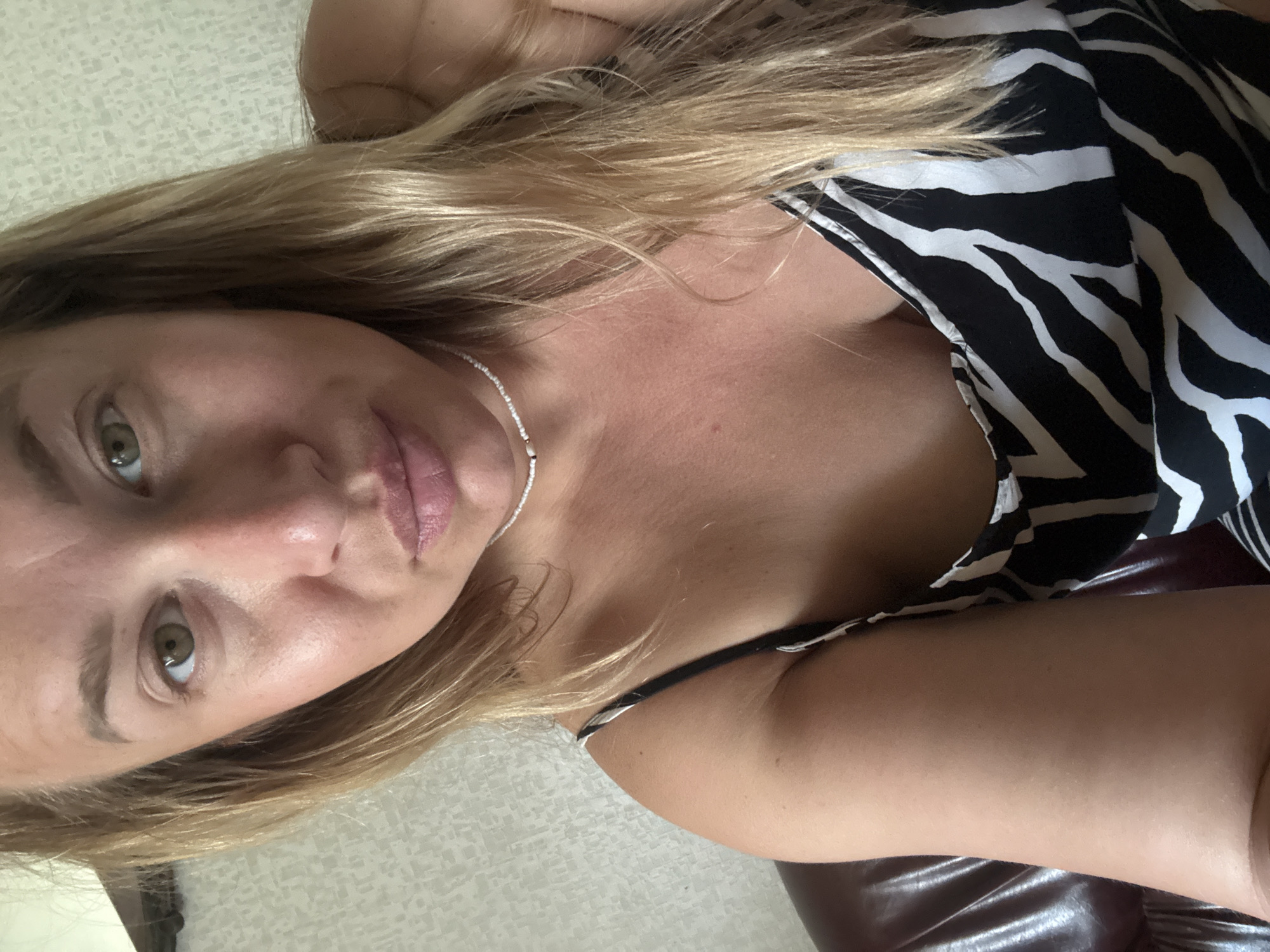 A close-up, waist-up, selfie-style photograph captures a woman with blonde hair and green eyes. She is wearing a black and white zebra-print camisole and a beaded necklace. The woman is seated on a dark brown leather surface, likely a sofa or chair. The background consists of a textured wallpaper. There is no indication of the city of Valday, Russia, in the image or its context.