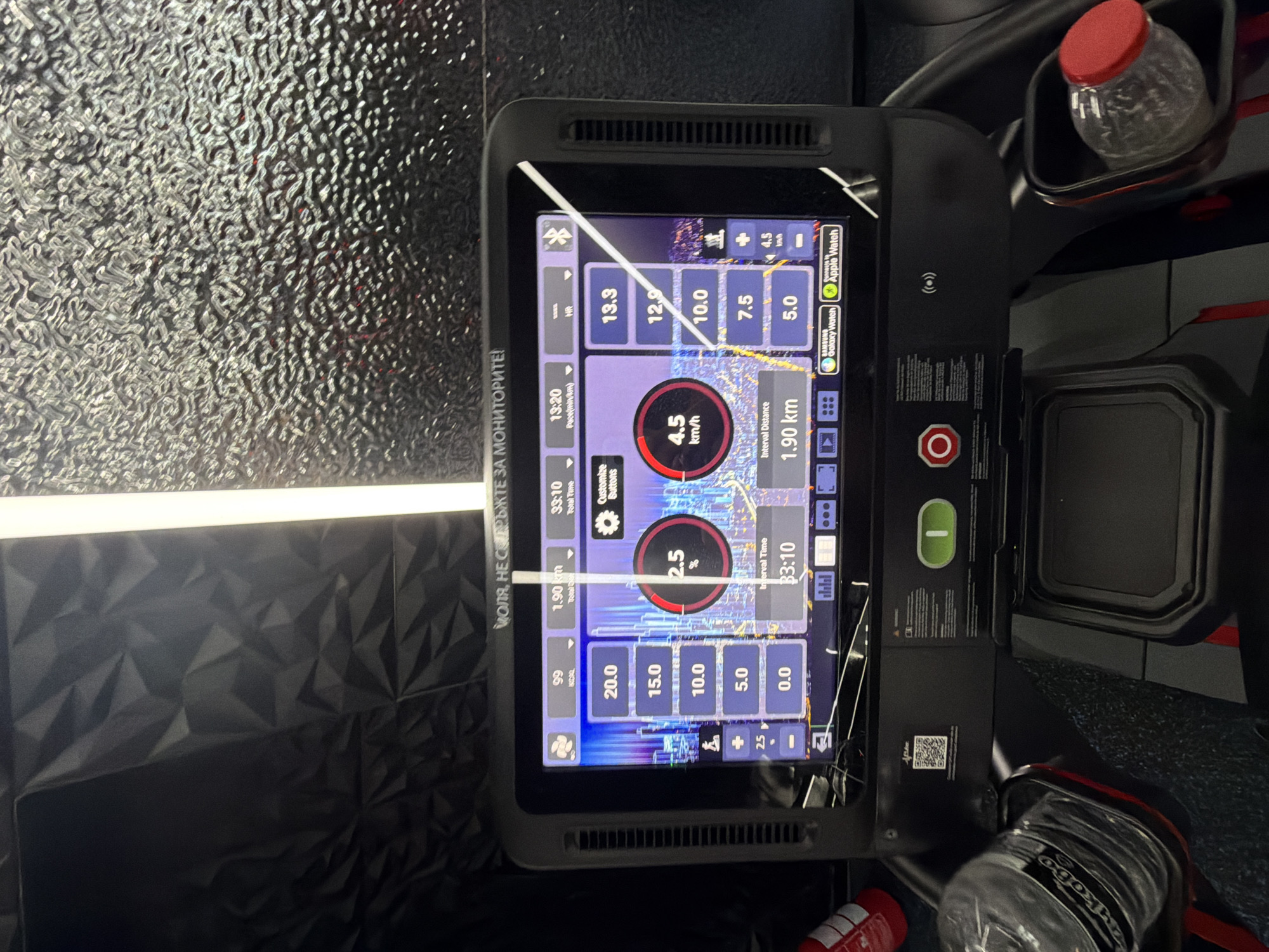 The image shows a close-up of a treadmill console in what appears to be a gym in Plovdiv, Bulgaria. The treadmill console has a large touchscreen display showing various workout metrics. On the screen, we can see values such as "99 kcal", "1.90 km", and "33:10 Time". There are also graphical representations of heart rate or intensity levels, showing percentages and speeds in km/h. Below the main display are buttons and indicators for workout controls, including an emergency stop button. To the left of the console, a water bottle is visible in a holder. To the right, another water bottle is partially visible. The background features textured wall panels and lighting. The overall environment suggests an indoor fitness facility.