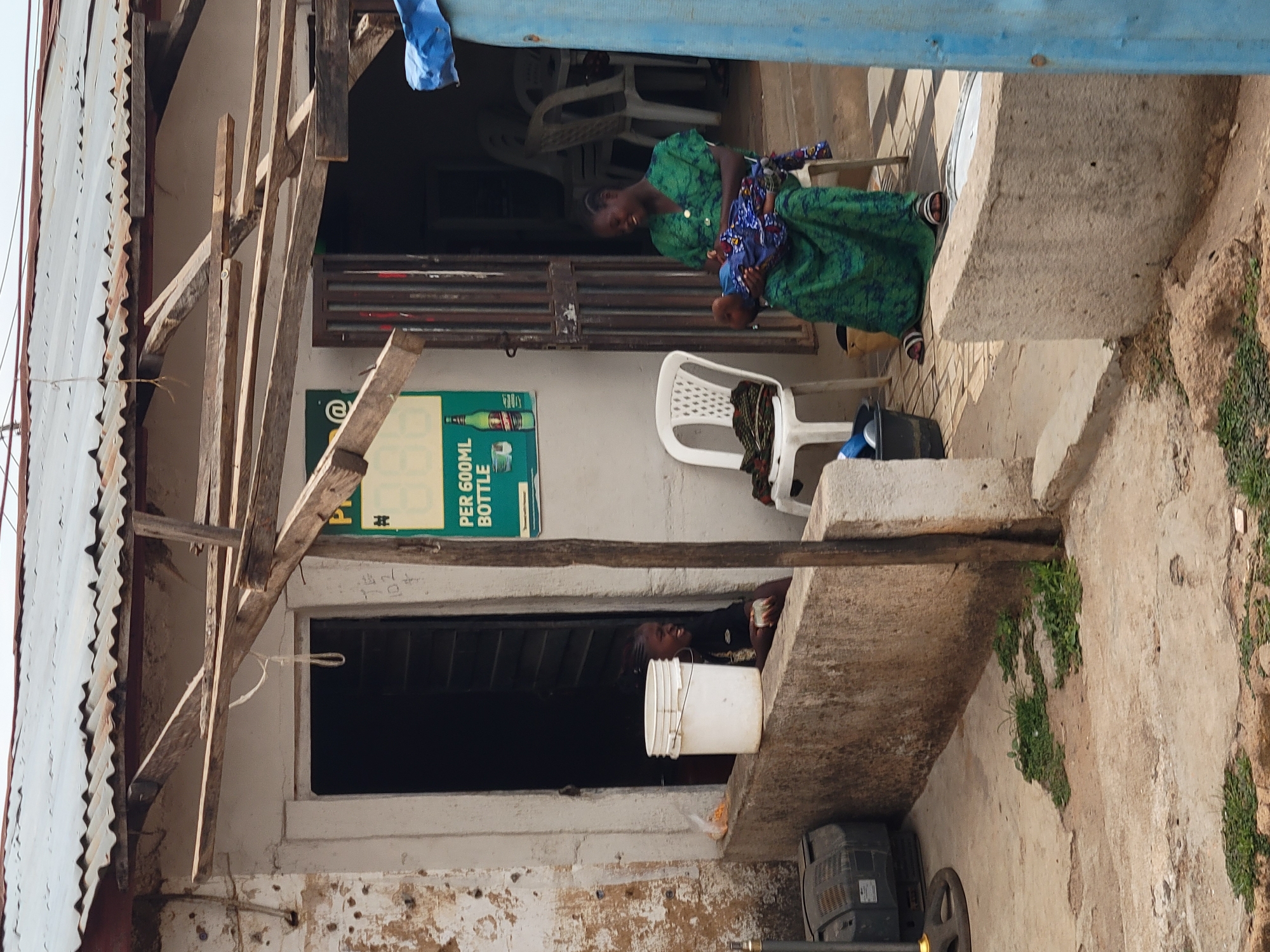The image depicts a woman in a green dress sitting outside a weathered building, holding a baby in her lap. The location is likely in Jos, Nigeria, based on the typical architecture and environment of the area.
The building shows signs of age and disrepair with a corrugated metal roof supported by exposed wooden beams. The walls are cracked and stained, with some vegetation growing on the exterior. A faded advertisement for a beverage is visible on the wall.
A second person is seen in the lower left of the image. She is partially obscured by the edge of the structure and appears to be sitting with a white bucket. There is an old television or electrical equipment visible near her.
The overall setting suggests a low-income, rural area, and the scene captures a moment of everyday life.