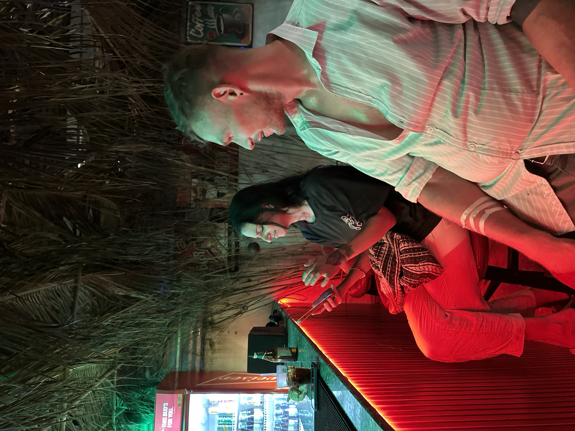 The image depicts two individuals at a bar or casual establishment, illuminated by prominent red and green artificial lighting, suggesting a vibrant nighttime atmosphere. The scene is oriented vertically, showing the subjects positioned alongside a long counter.
To the right, a man with short, light-colored hair and a light beard wears a striped, unbuttoned long-sleeved shirt. He is looking downwards and slightly to his left, seemingly engaged with the person next to him. Red and green lights cast strong, colored shadows and highlights across his face and clothing, and a white band is visible on his left forearm.
Beside him, a woman with long dark hair is holding a smartphone, her attention focused on the device. She wears a dark t-shirt and has a patterned cloth draped over her lap. Her posture suggests she is either seated or leaning against the counter.
The counter in front of them features a strong red LED strip along its edge. On the counter, a glass containing a drink and a bottle are visible, along with a dark speaker or box. Below the counter, a large red cooler displays the text "Budweiser" vertically and the slogan "THIS BUD'S FOR YOU."
The background is characterized by dried palm fronds or similar tropical foliage, creating a rustic and natural texture behind the bar. Above the man's head, a dark sign with white text reads "Coffee" and features a coffee cup icon. The overall ambiance is relaxed and casual, indicative of a tropical or beachside bar setting, such as in Mũi Né, Vietnam.