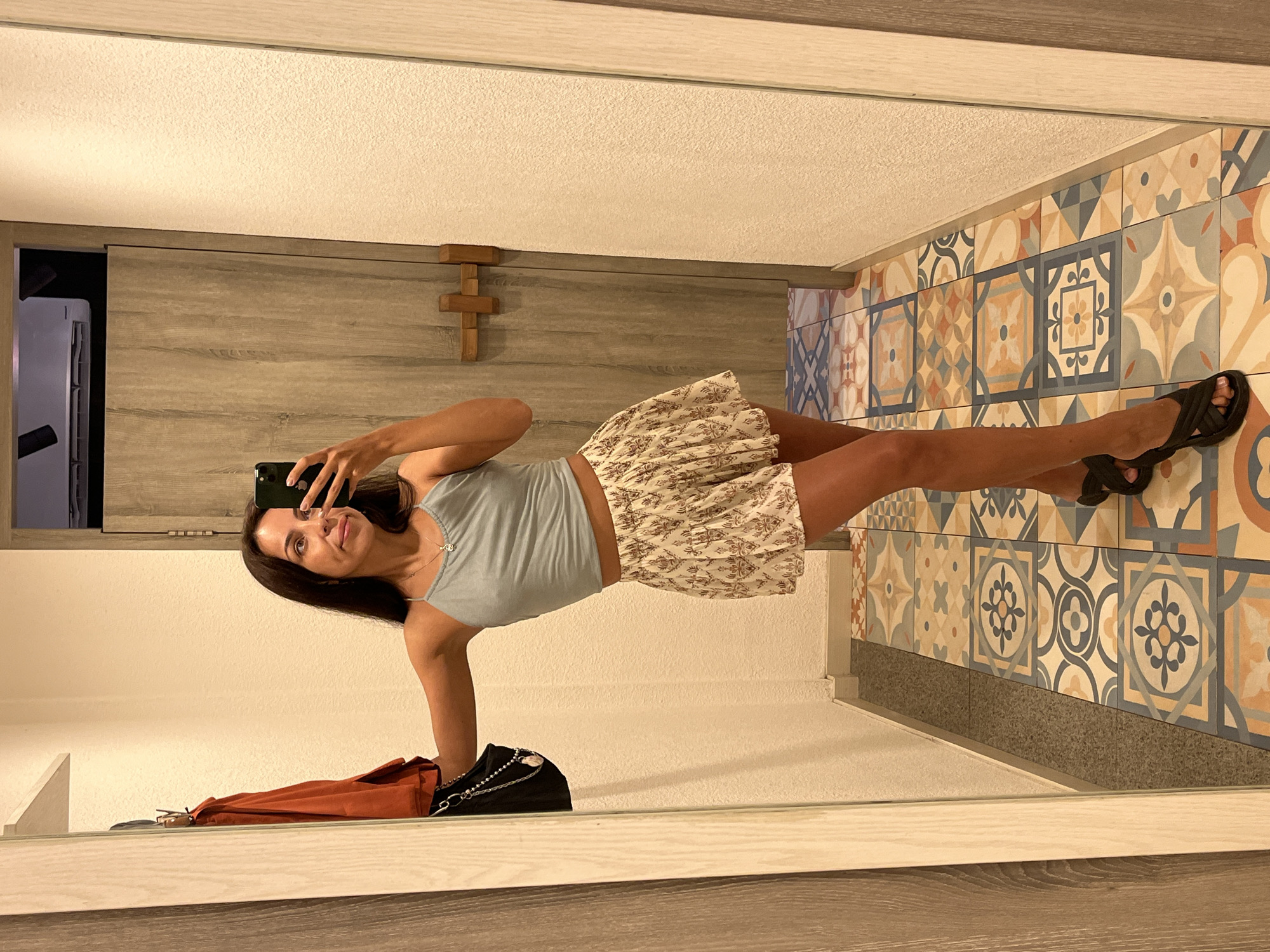 This image captures a mirror selfie of a young woman, presented with a 90-degree counter-clockwise rotation. The woman, who has long dark hair, is reflected standing upright, holding a dark smartphone in her right hand to take the photo. She is wearing a light blue crop top, a short skirt with a floral or intricate pattern in light colors, and dark sandals. A small pendant necklace adorns her neck, and she displays a soft, pleasant smile while looking towards her phone screen.
The setting appears to be an indoor space, potentially a room or hallway, with the provided context indicating Kuta Selatan, Indonesia. To the right of the woman, reflected as a wall, are decorative square tiles featuring geometric and floral motifs in shades of blue, orange, beige, and gray, with a dark gray trim along the bottom. Behind her, a plain light-colored wall is visible. To her left, a wooden sliding door or panel is reflected, above which hangs a small, wooden, cross-like decoration. A partially visible white appliance is seen to the far left. On a surface in the foreground, reflected in the mirror, are a dark bag with a chain strap and an orange-brown item, possibly a folded piece of clothing or another bag. The ceiling is white and textured. The lighting is artificial and bright, illuminating the scene clearly. There is no visible text in the image.