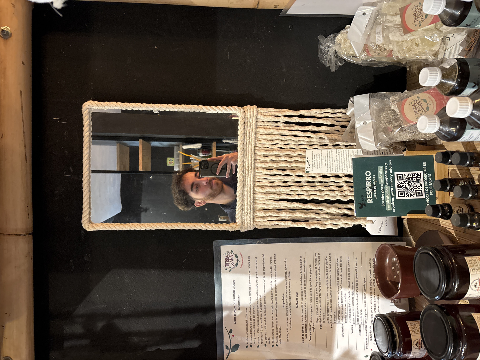 Here is a concise description of the image:
The image is taken from an overhead perspective on a table in Puebla, Mexico. A mirror framed in rope sits on the table, reflecting a man holding a smartphone, presumably taking the photo. Next to the mirror are bottles and jars of natural products. A product information card with the "TIERRA SANA" logo is also present, as well as a card advertising "RESPIRRO" jarabe de Maguey. The table itself is a dark color, and a bamboo-like structure is visible along the left edge of the frame.