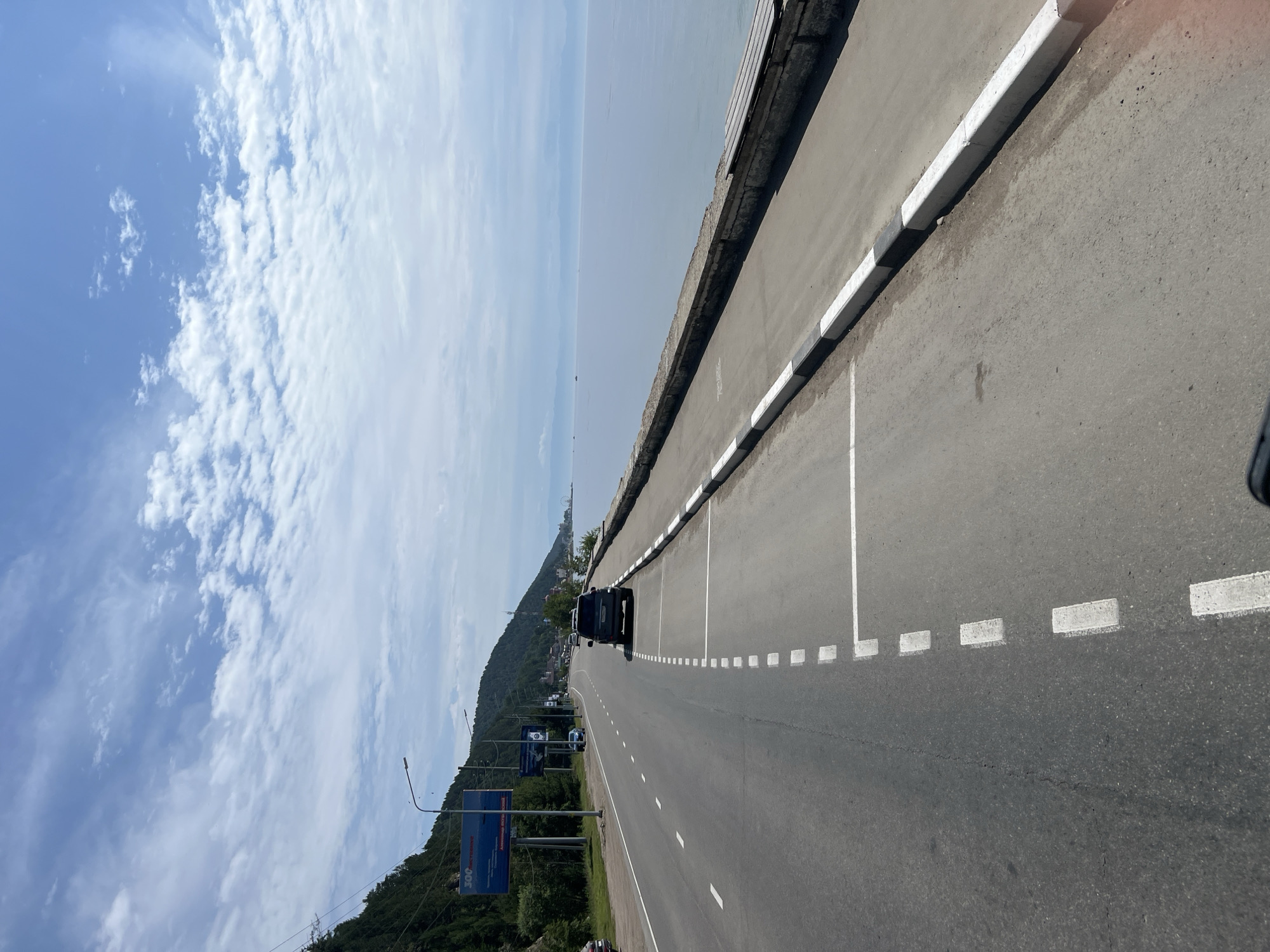 The image captures a scenic view along a road beside a large body of water, likely Lake Baikal, in Listvyanka, Russia. A paved road runs parallel to the water's edge, with a distinct concrete embankment separating the road from the lake. White dashed lines mark parking spaces along the roadside. A dark-colored SUV is parked in one of these spaces. In the distance, the road curves and is lined with trees and buildings, suggesting a town or settlement. The sky is a bright blue with scattered white clouds, indicating fair weather. A few boats are visible on the calm water. The overall scene depicts a well-maintained road with infrastructure for parking and a pleasant natural environment.