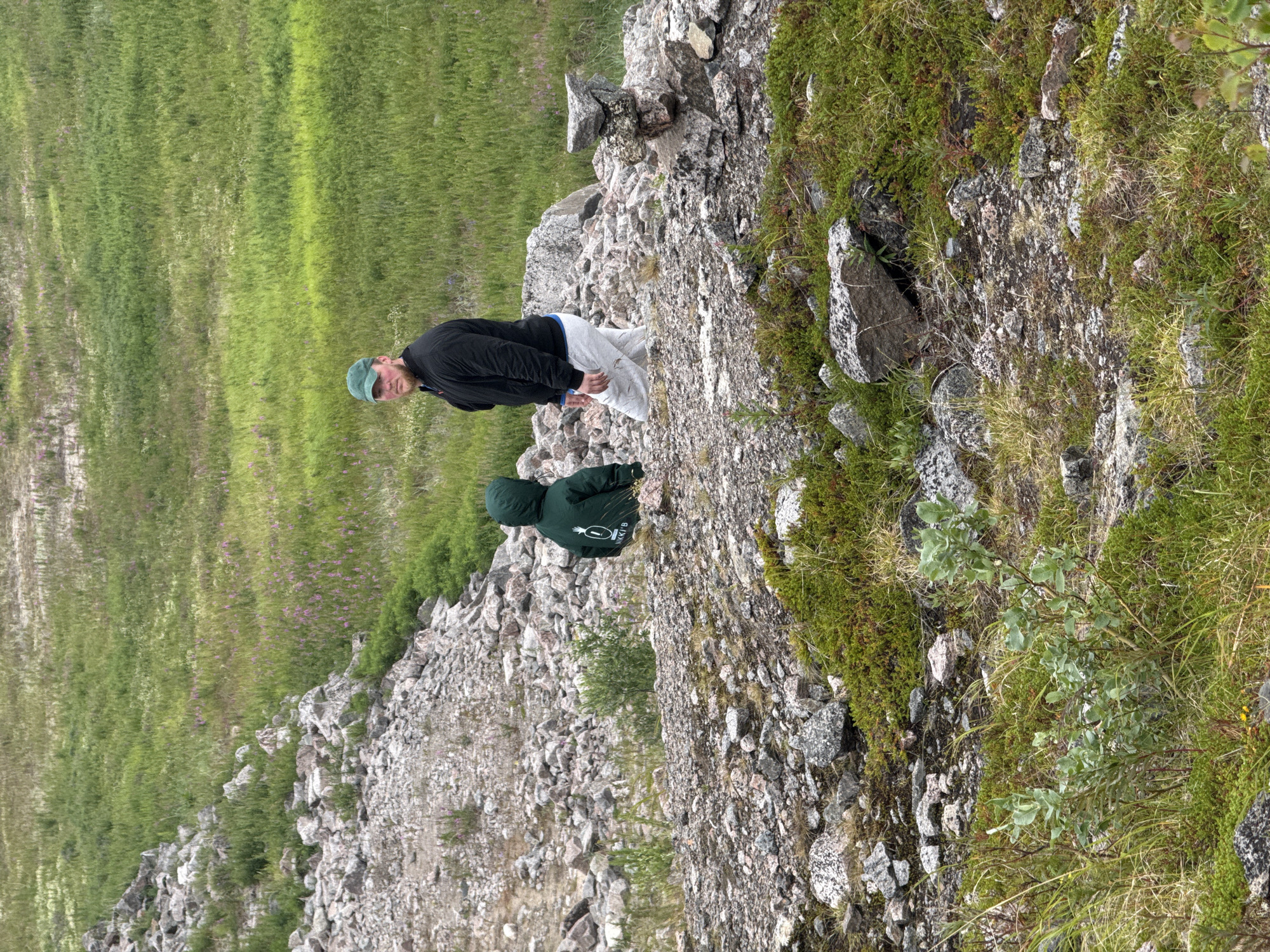 Two individuals are positioned in a rocky, vegetated terrain. A person wearing a green hooded jacket is facing away from the camera, seated on the ground. To their right, a man in a black jacket and light-colored pants, wearing a green baseball cap, is standing and looking towards the camera. The background consists of a green, grassy expanse dotted with purple flowers and a slope of grey rocks. This appears to be an outdoor scene in a natural environment, possibly on the outskirts of Teriberka, Russia, given the rugged landscape.