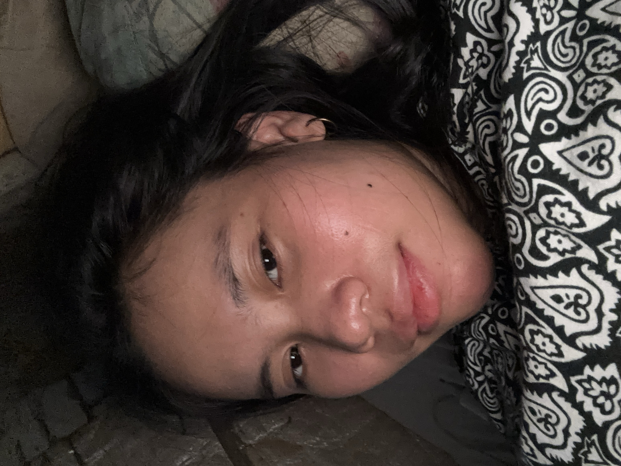 A close-up selfie of a young woman is captured from a slightly high angle. She is lying down, as indicated by her hair spread out and the pillow beneath her head. Her eyes are open, and she is looking towards the camera with a soft smile. Her skin appears slightly flushed, and there are a few small moles visible on her face. She is wearing a simple hoop earring in her left ear and a black and white patterned garment, likely a shirt or blanket, is visible around her neck and shoulders. The background appears to be indoors, with a dimly lit, textured surface on the left, possibly a wall or rough bedding, and a patterned textile on the right. The lighting suggests it might be nighttime or a dimly lit room. The image does not contain any specific elements that would indicate its location within East Jakarta, Indonesia.