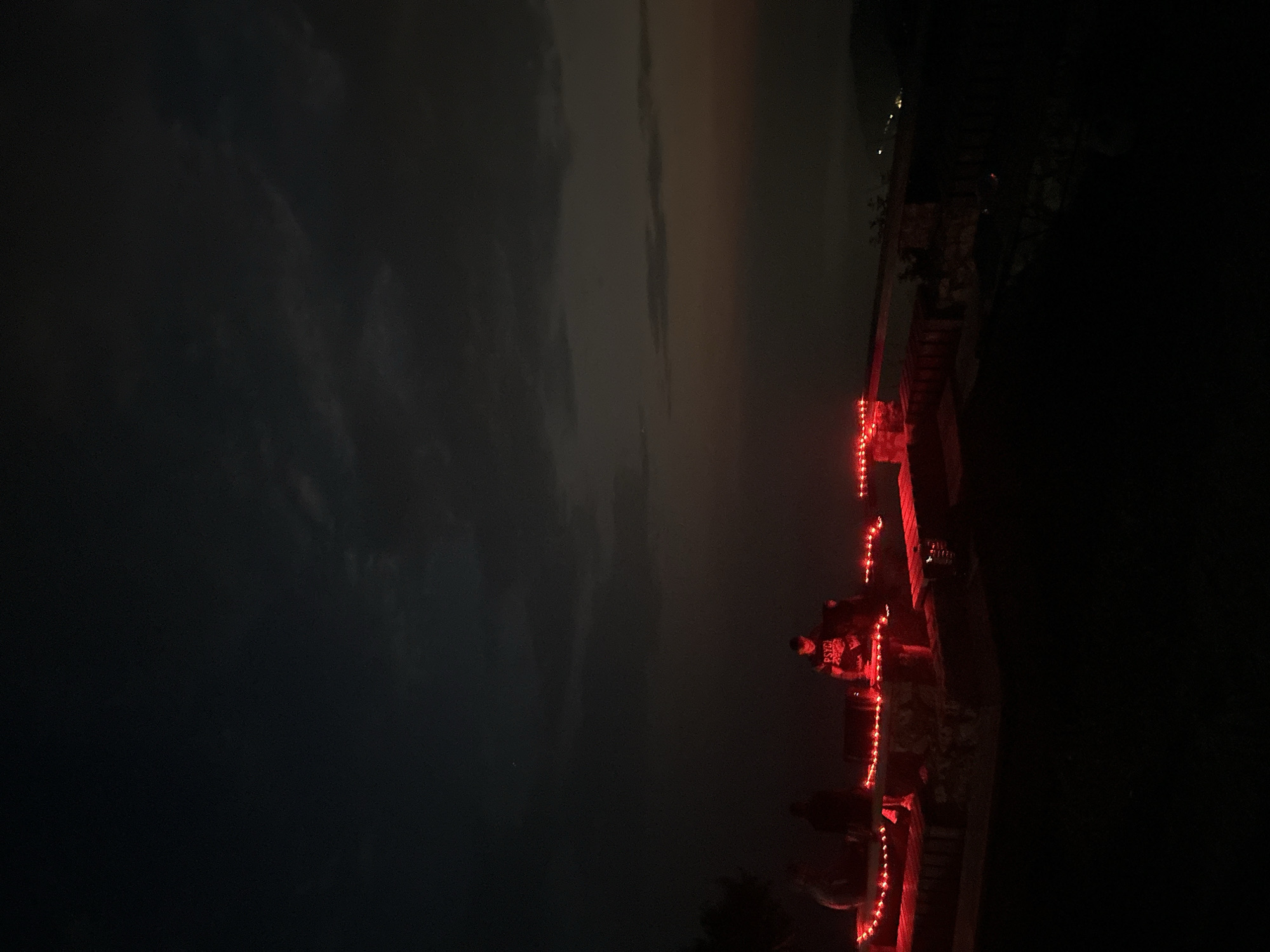 In the city of Gluhi Do, Montenegro, at dusk, a group of individuals are gathered on a raised platform illuminated by strings of red lights. One person in the center appears to be operating a DJ setup, with the word "PSYCH" visible on their shirt. Two other individuals are seated to the left of the DJ. The platform is made of stone and wood, with benches. In the background, a hazy vista of distant lights suggests a city or town below, seen through the twilight sky. The overall scene conveys a sense of a nighttime gathering or event.