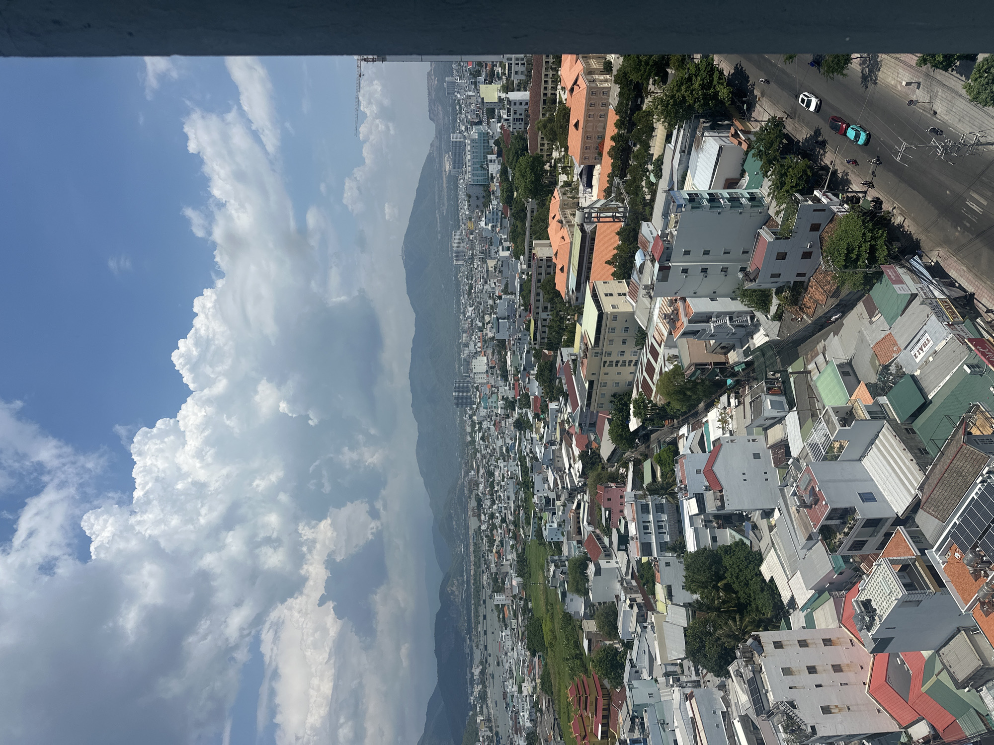The image presents a high-angle, panoramic view of a densely populated urban area in Nam Nha Trang Ward, Vietnam, under a partly cloudy sky. The foreground is dominated by the edge of a tall building, rendered in a neutral gray tone. The mid-ground reveals a sprawling cityscape characterized by a mix of low-rise and multi-story buildings, many with distinctive orange-tiled roofs. Residential structures, businesses, and some larger institutional buildings are visible. Lush green trees and vegetation are interspersed throughout the urban landscape, particularly in courtyards and along street edges.
In the background, a hazy mountainous terrain forms the backdrop to the city. A body of water, likely a bay or river, is visible in the distance, dotted with boats. The sky is a vibrant blue, with large, billowy white and gray cumulus clouds creating dynamic shadows and highlights across the scene. A construction crane is visible on the right side of the image, indicating ongoing development.
The streets in the city are paved and appear to be active. Several vehicles, including cars and motorcycles, are depicted traversing the roads. Pedestrians are also present, though their details are indistinct due to the distance. The overall impression is one of a bustling, tropical city with a blend of modern and traditional architecture, set against a natural landscape.