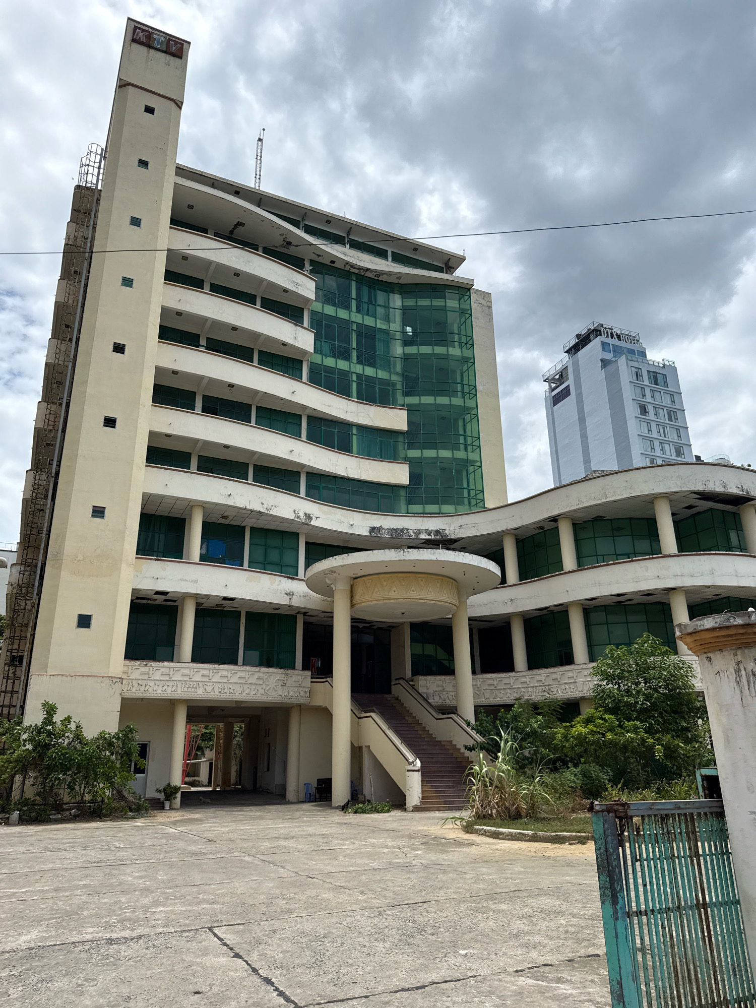 The image shows a large, light-colored, multi-story building in Nha Trang, Vietnam. The building has a modern architectural style with curved balconies and large green-tinted windows. A tall, slender tower with a "KTV" sign is attached to the left side of the building. The building appears to be unoccupied or in a state of disrepair, with some visible signs of weathering and peeling paint. The foreground consists of a concrete courtyard and a patch of overgrown vegetation. In the background, another taller, white building with a "DTX HOTEL" sign is visible. The sky is overcast with gray clouds.