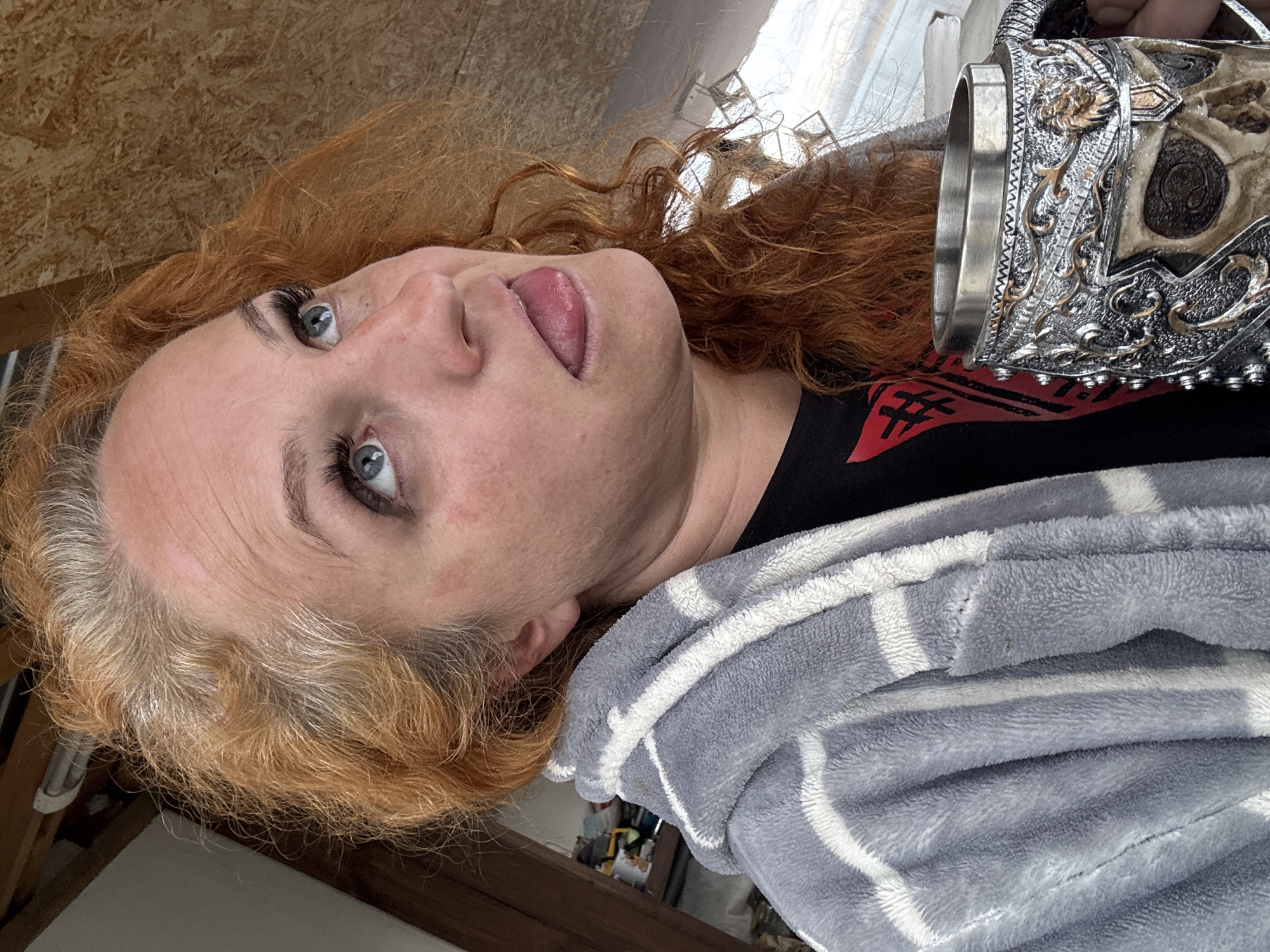 The image features a close-up, rotated portrait of a woman with vibrant red, curly hair, which shows natural lighter/grey regrowth at the roots. She has fair skin and striking blue eyes, enhanced by mascara. Her tongue is playfully extended as she looks upwards and to her left. She is dressed in a black top with a partially visible red graphic design and is covered by a soft, grey blanket or robe with white stripes.
In her hand, she holds an ornate metal mug or goblet. This object is highly detailed with silver-colored embossed patterns, gold accents, and incorporates a skull or skeletal face design. The scene is indoors, brightly lit by ambient light, suggesting daytime. A textured light brown wall, possibly made of OSB or plywood, is visible in the upper background, indicating a casual or rustic indoor setting. The woman's expression and attire convey a relaxed, comfortable, and perhaps quirky mood. The red graphic on her shirt is abstract and not clearly decipherable. The context for the photo is Chaikovsky, Russia.
