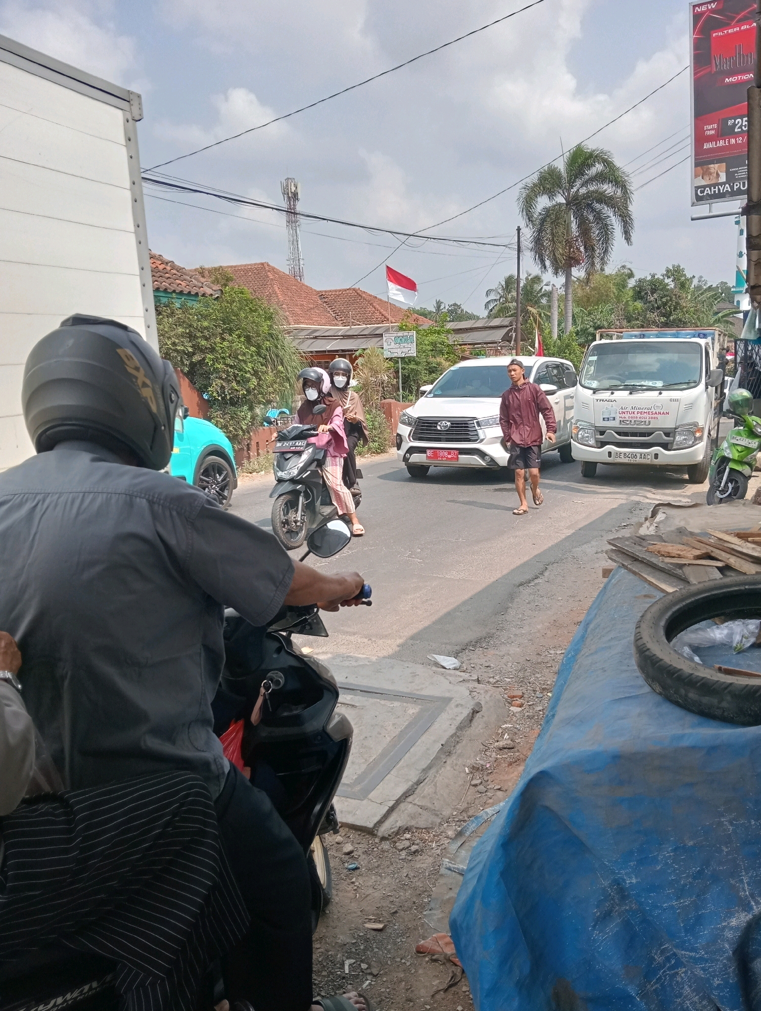A man in a black helmet and gray shirt rides a motorcycle, with a portion of his striped pants visible. He is in the foreground, facing away from the camera. To his right, on the road, a white SUV is parked, and a young man in a maroon jacket and black shorts walks across the street. Behind the SUV, a white truck is also parked. Further down the road, two people on a black scooter are visible, the passenger wearing a light pink shirt. A turquoise car is parked on the left side of the road. The scene appears to be a street in a town or city, with buildings and trees lining the road. The sky is partly cloudy. There is a large blue tarp covering something on the right side of the image, with a tire resting on top of it. A billboard with a cigarette advertisement is visible in the background.