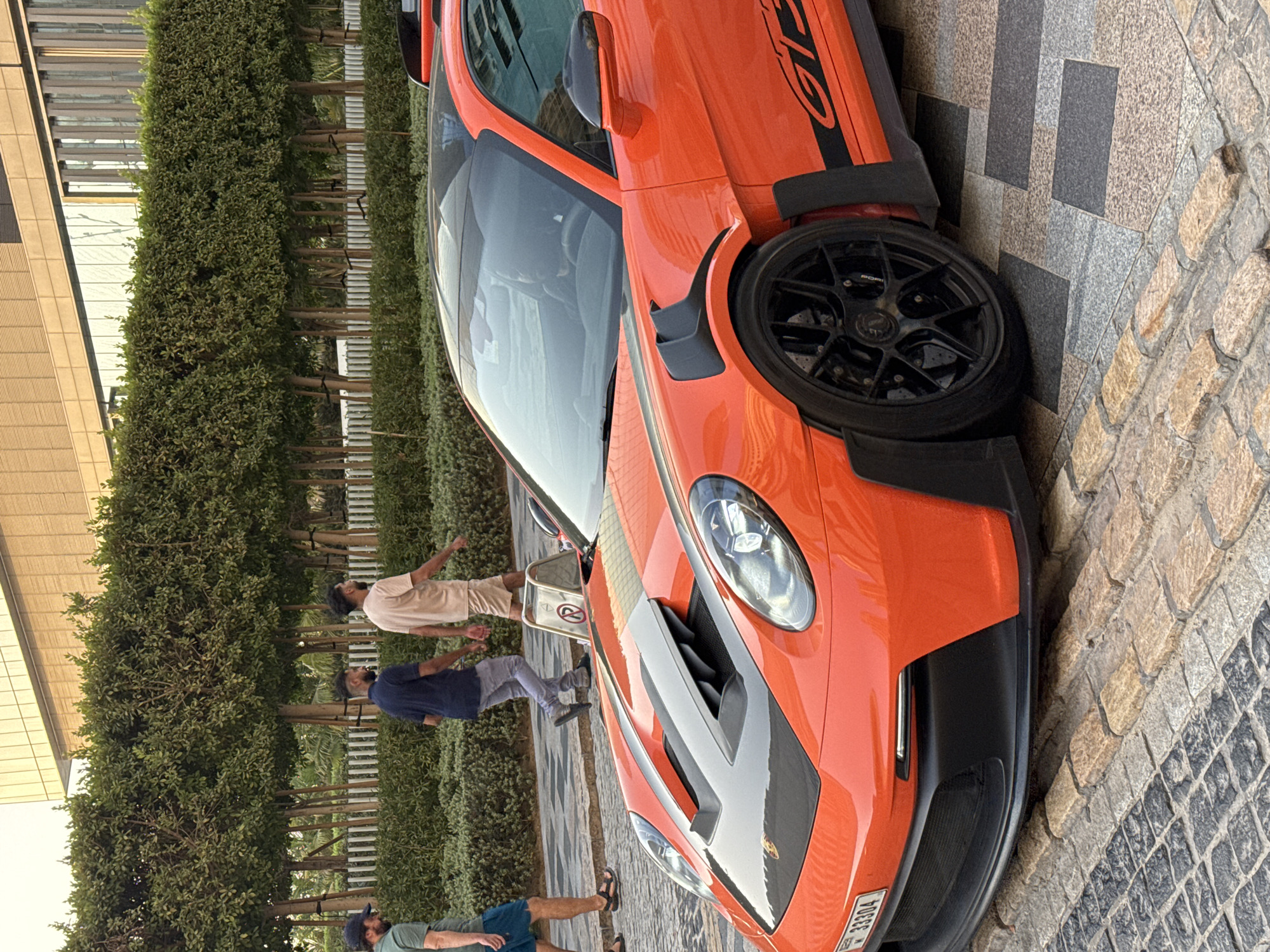 A bright orange Porsche GT3 RS is parked on a cobblestone pathway in Dubai, United Arab Emirates. The car is the primary subject, with its distinctive aggressive styling including large air vents on the hood and fender flares. The vehicle features black racing stripes along its side and black wheels. Three men are walking in the background, appearing to be in motion. One man on the far left wears a green t-shirt and blue shorts. Another man in the middle wears a dark blue t-shirt and grey pants. The third man on the right wears a light-colored t-shirt and shorts. Behind the men, a row of neatly trimmed green bushes and trees form a natural hedge, separating the pathway from a modern building. The building has a glass facade and is illuminated by natural light. The sky is overcast, suggesting it is late afternoon or early morning. The overall scene depicts a moment in an urban environment with a focus on a high-performance automobile.