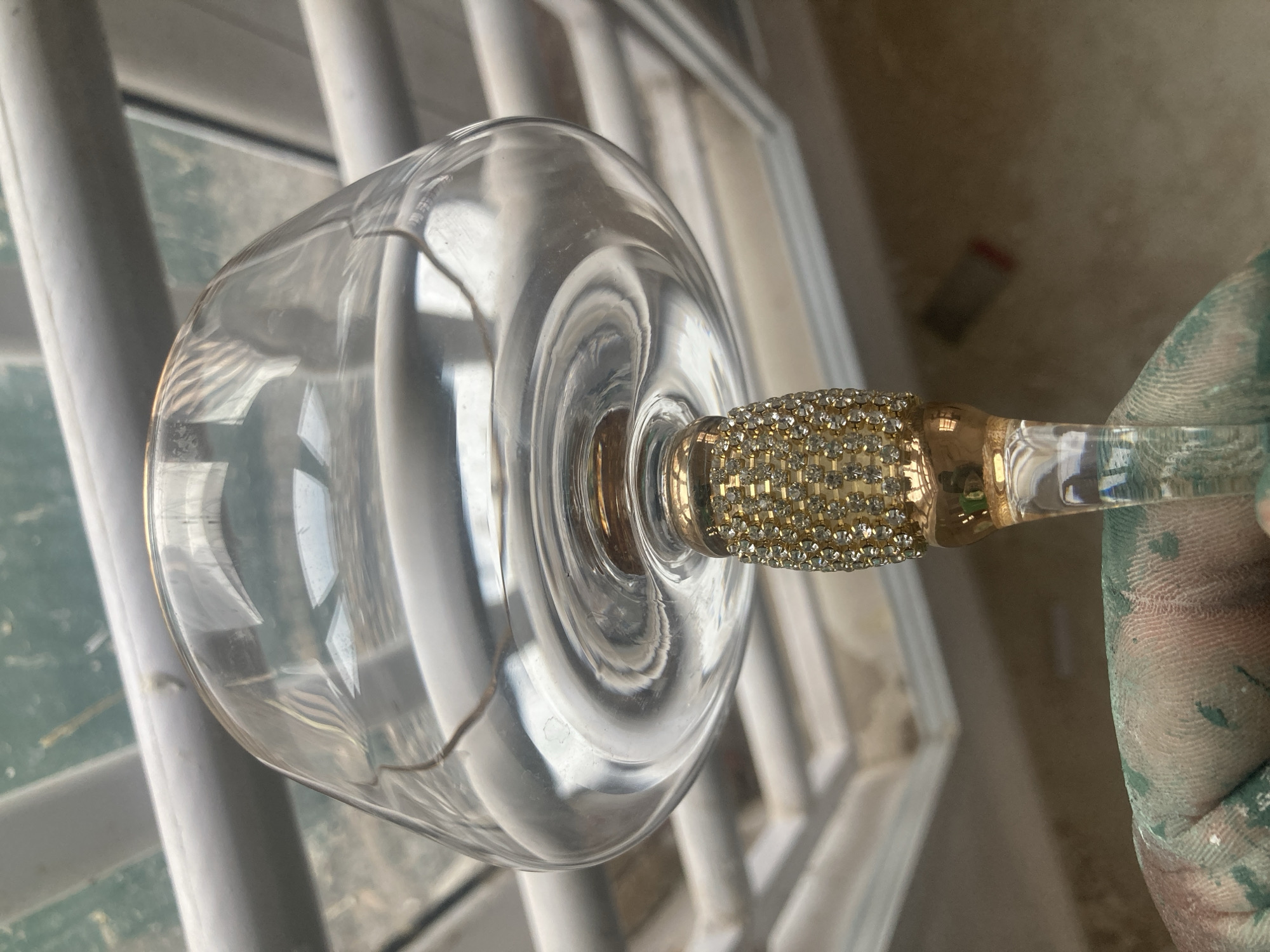 A clear glass wine glass with a gold-colored stem adorned with rhinestones is held by a hand with splatters of green paint. The glass is positioned in front of a white-painted railing. The location appears to be indoors, possibly a residence, as suggested by the railing and a glimpse of a room in the background. There is no discernible information that specifies the location to be Jos, Nigeria.